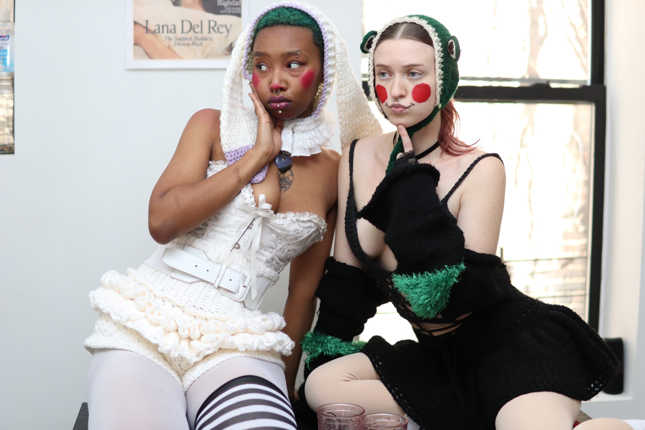 Two young women dressed in whimsical costumes posing together indoors near a window with a poster in the background. One is wearing a white dress with a ruffled skirt, striped tights, and a colorful crochet hood; the other is in a black crochet dress