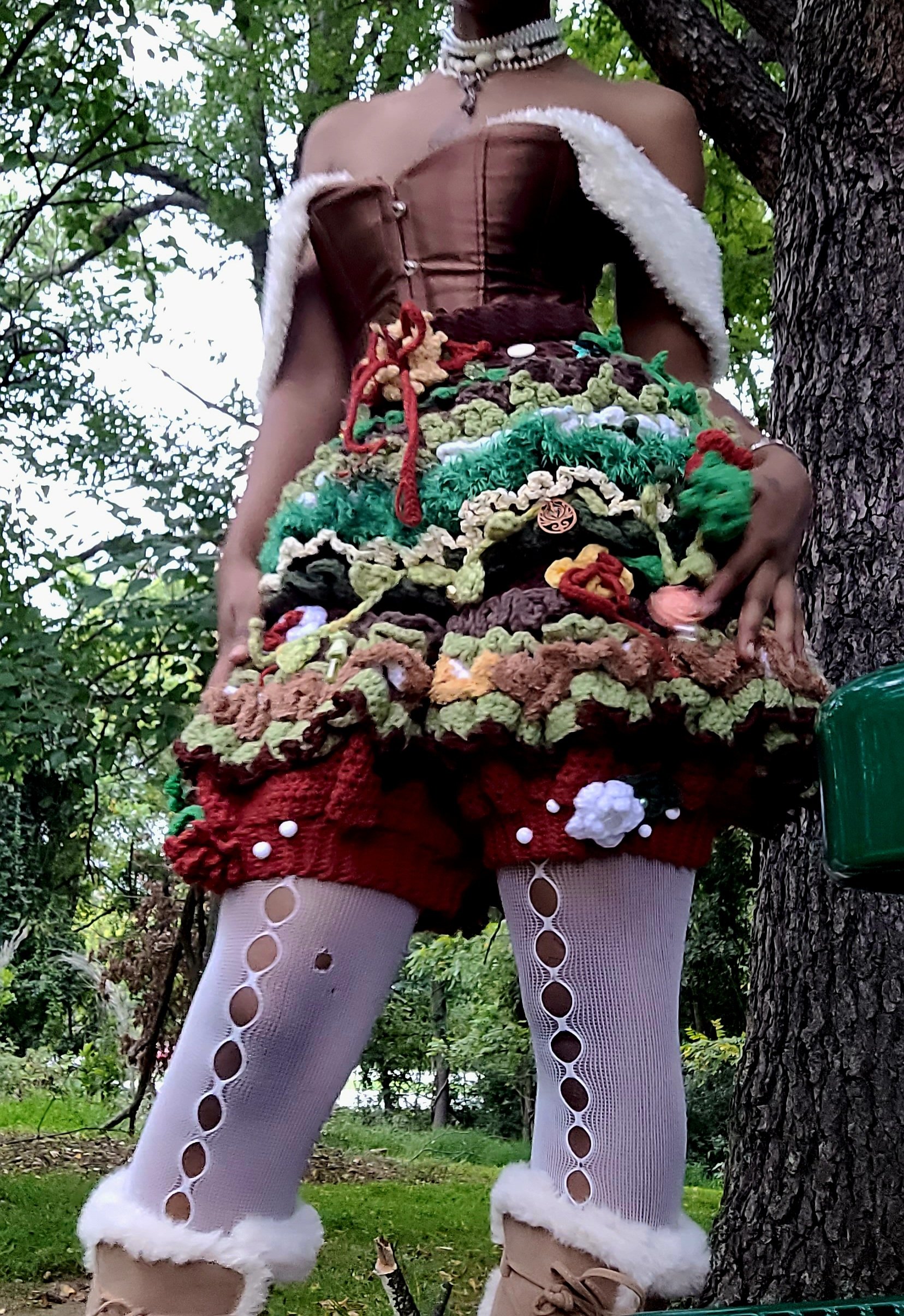 Person wearing a brown corset top, multicolored handmade or knitted outerwear, white tights with circular cutouts and fur trim, standing outdoors near a tree.