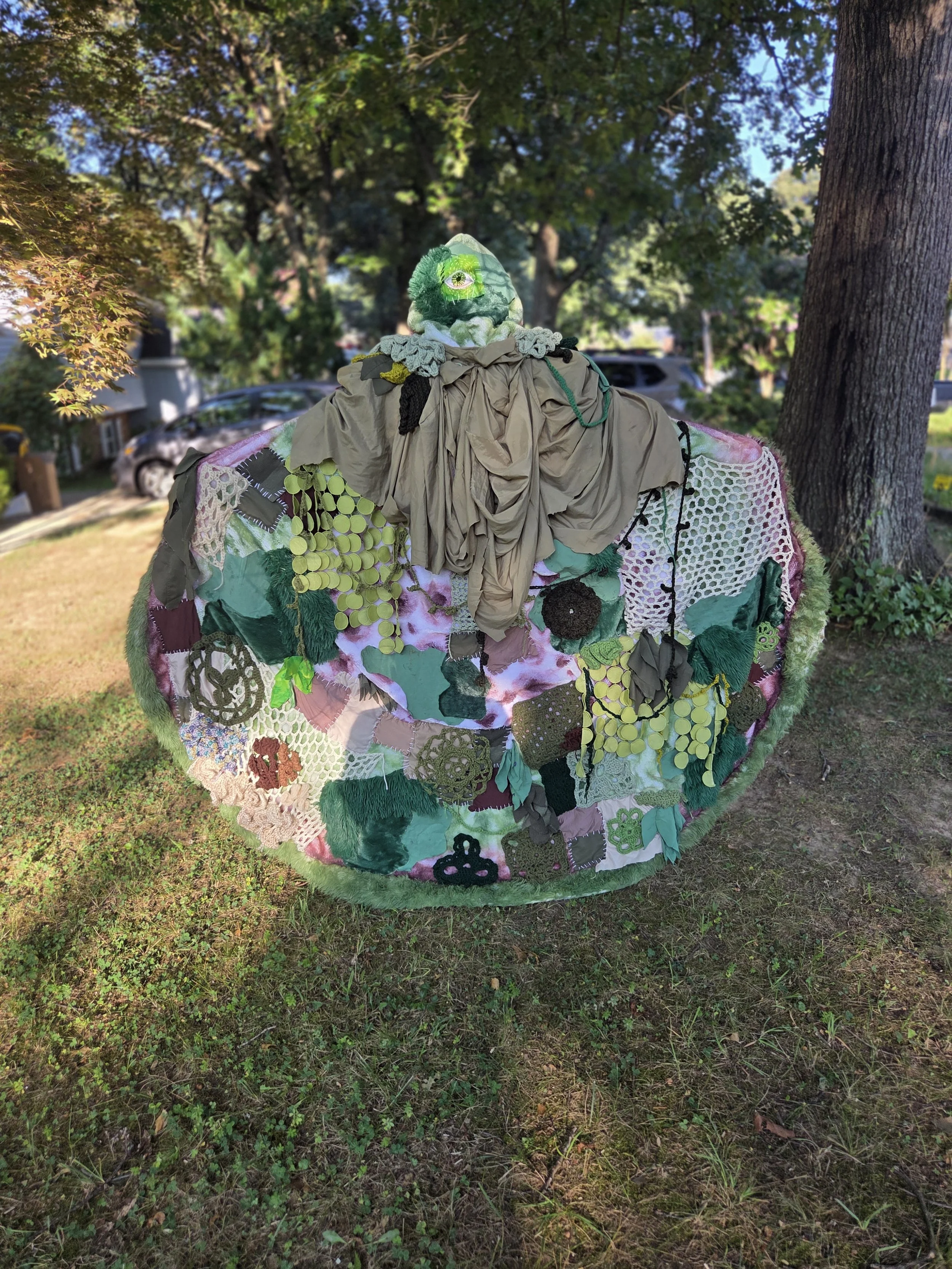A large painted sculpture resembling a turtle or tortoise, decorated with colorful fabric patches and textiles, situated outdoors on grass near trees in a residential neighborhood.