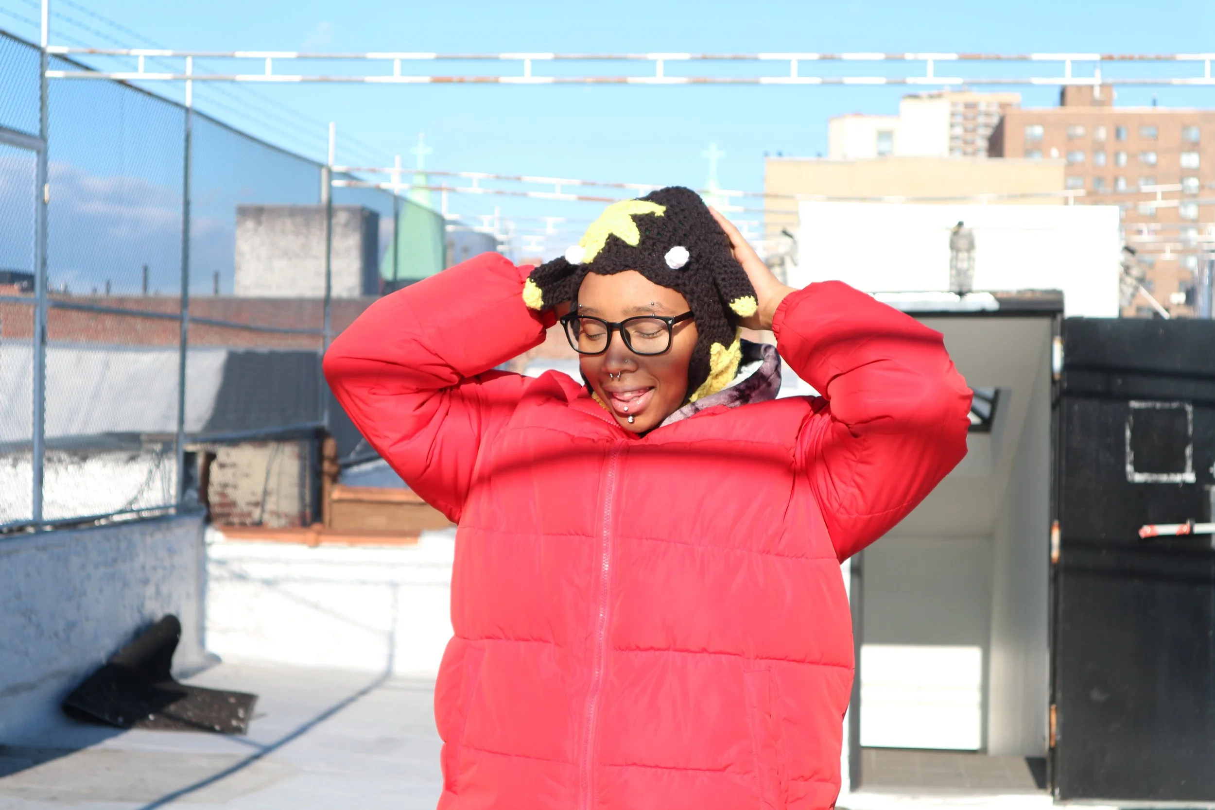 A person with glasses and piercings wearing a red jacket on a rooftop with a cityscape background, holding a plush hat on their head.