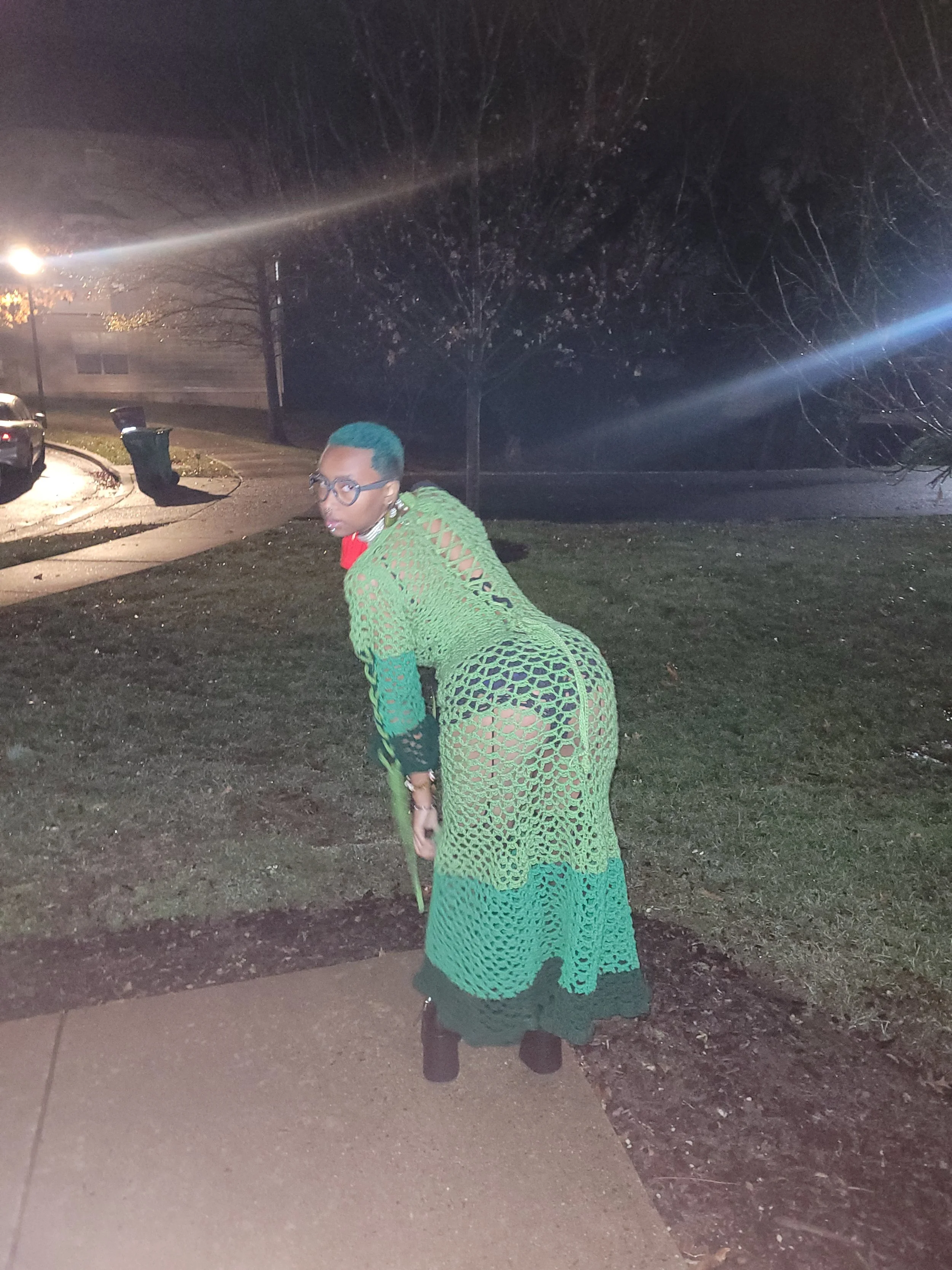 Person with teal hair, glasses, and green crochet dress outdoors at night
