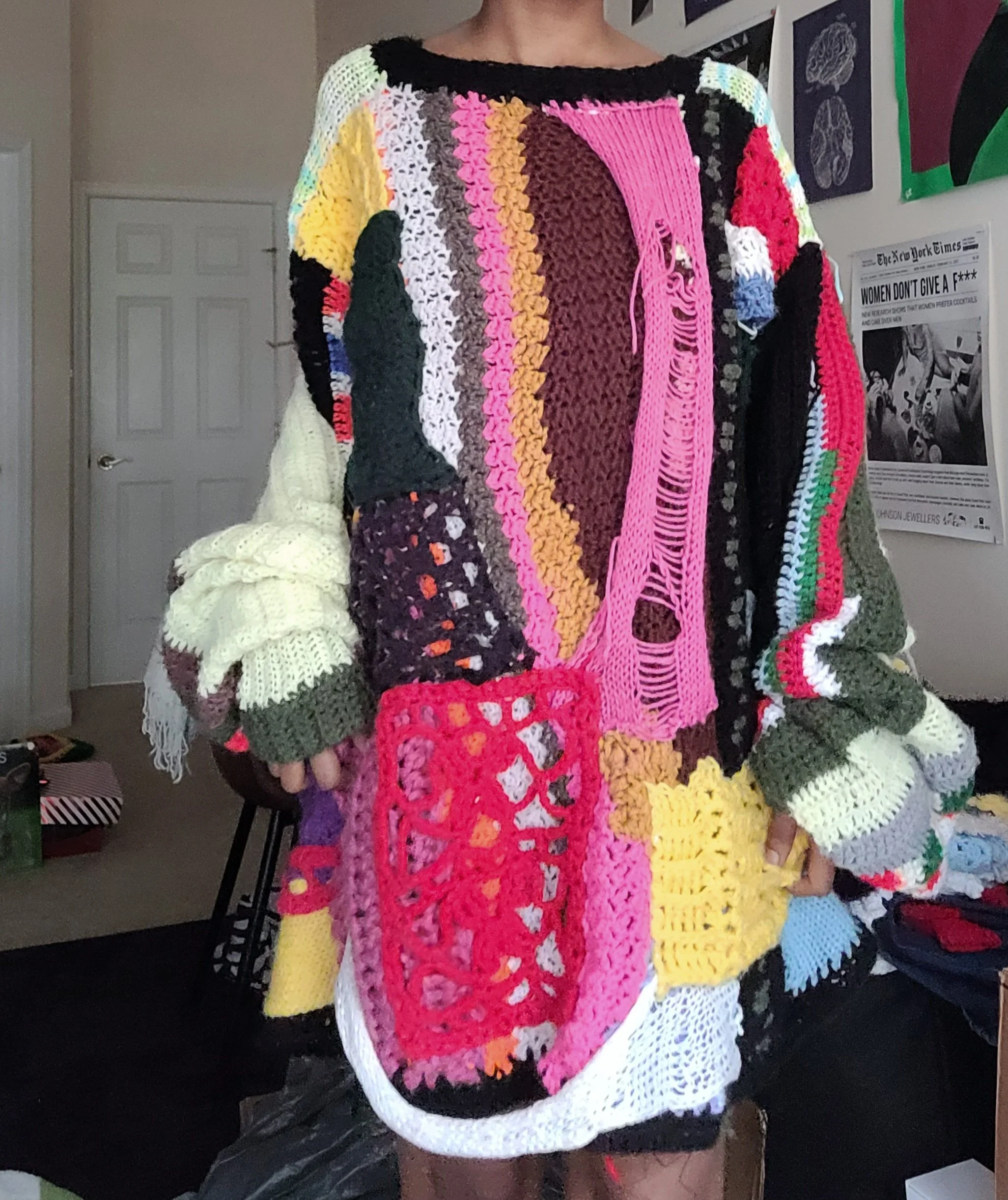 A person wearing an oversized, multicolored, patchwork crochet sweater with various patterns and textures.