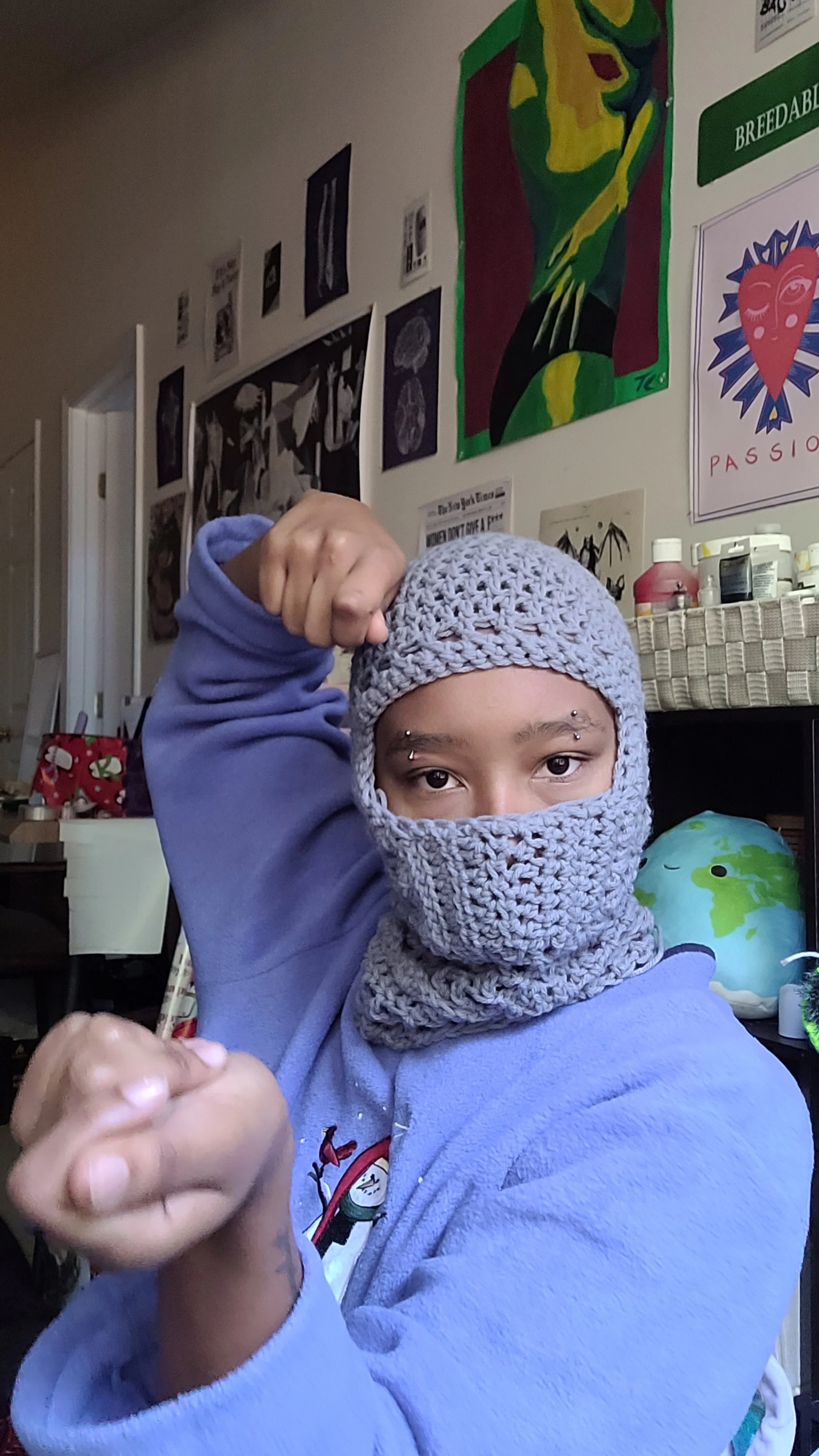 A young person wearing a lavender hoodie and a crochet face covering, with eyebrow piercings, flexes their arm. The background has colorful artwork and posters on the wall, with a globe pillow and various objects on a piece of furniture.