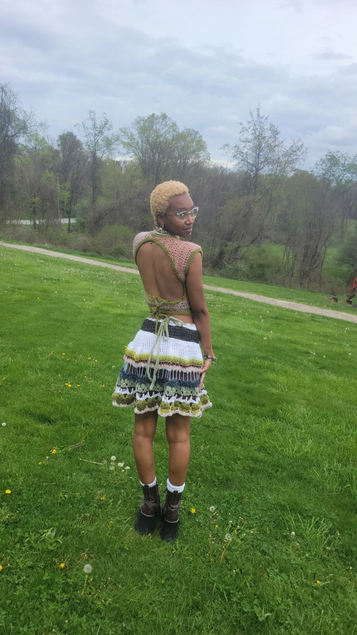 A woman with short blond hair, glasses, and dark skin standing outdoors on a grassy field with trees and cloudy sky in the background. She is wearing a colorful crocheted outfit with an open back, a multi-colored skirt, and brown boots.
