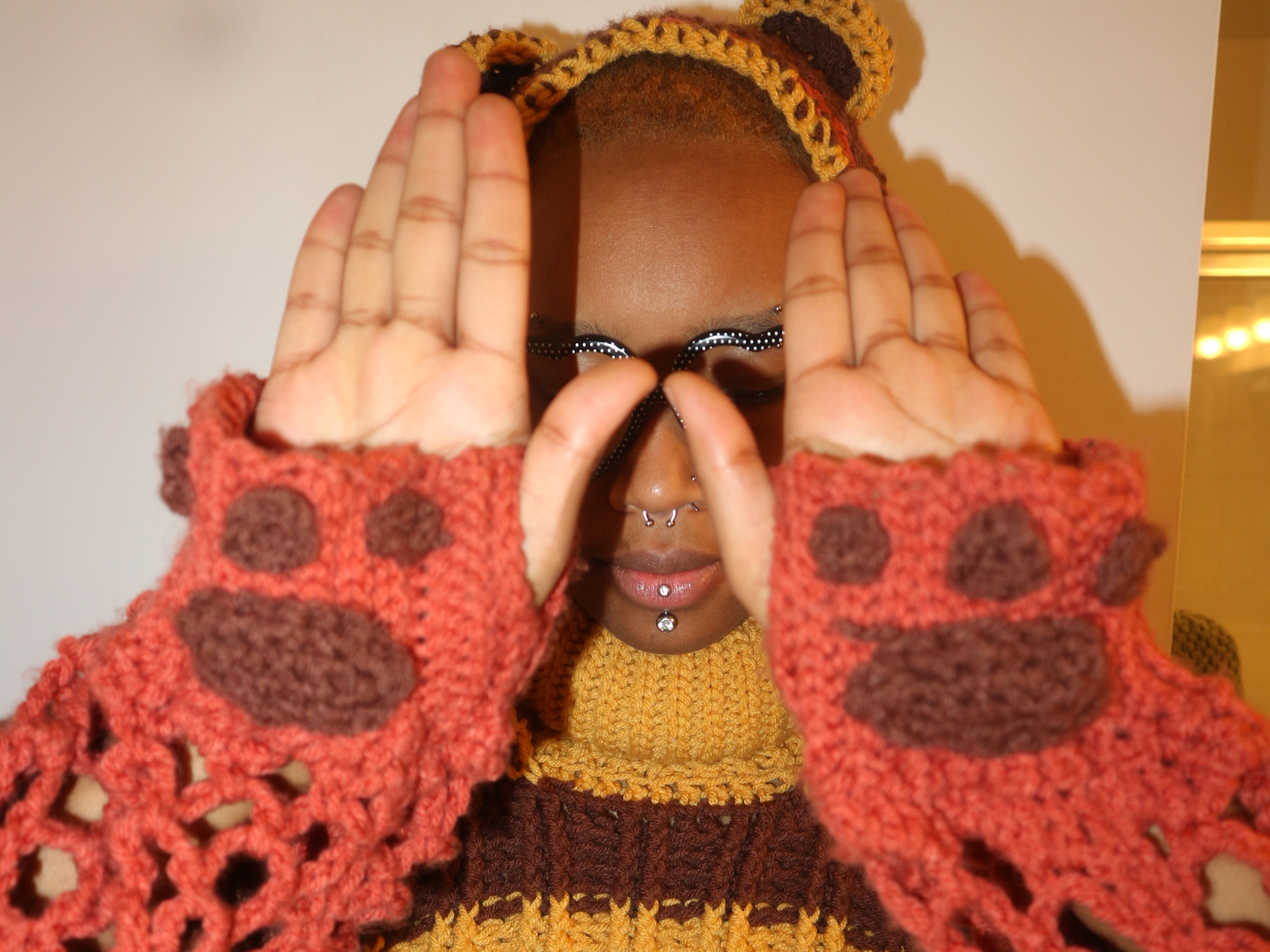 Person with glasses and colorful knitting sweater, holding hands up in front of face with fingers spread, wearing a multicolored knitted headband.