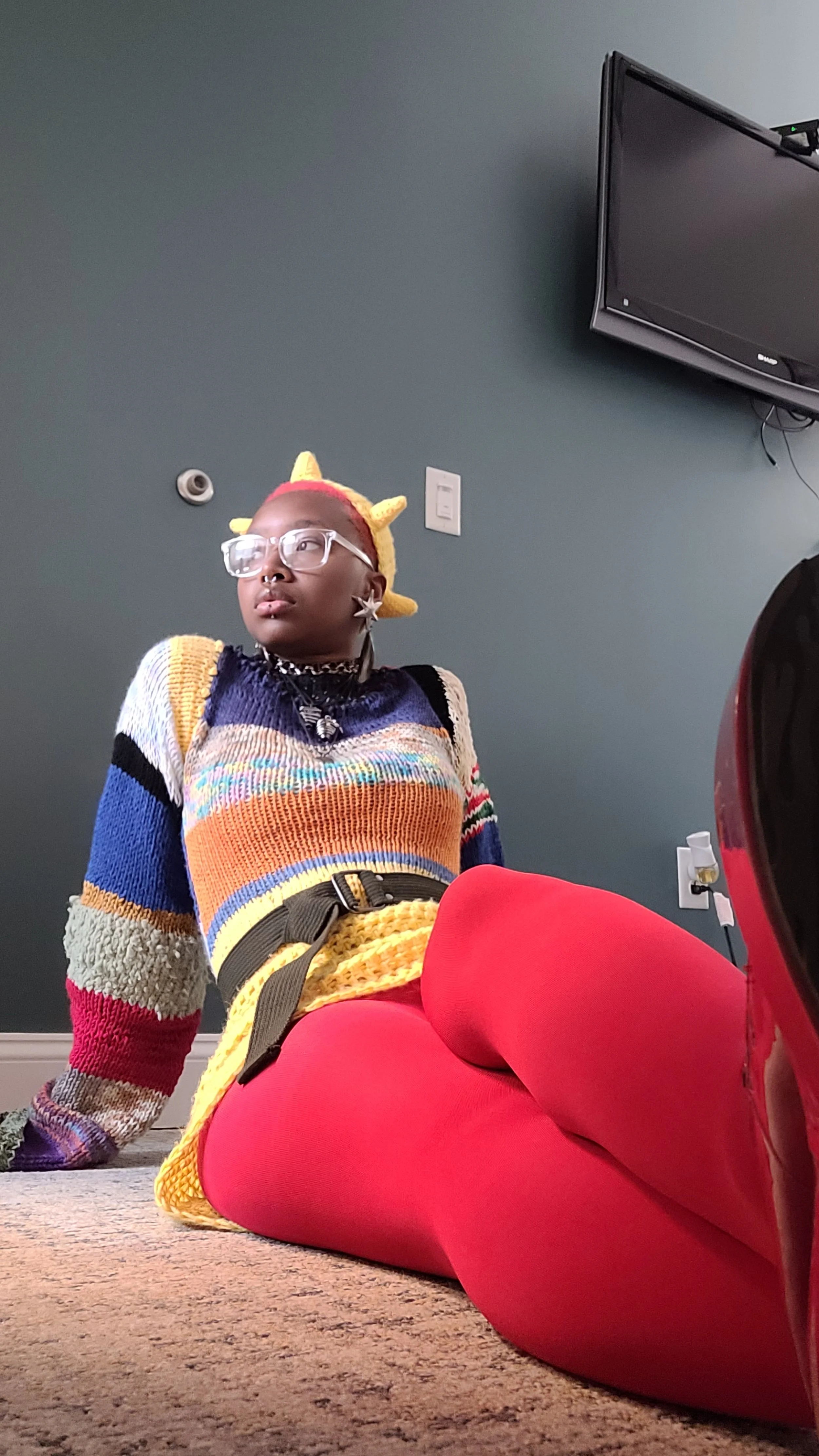 A person sitting on the floor wearing a colorful sweater, red tights, and a yellow wig with unicorn horns, with a wall, a television, and a power outlet in the background.