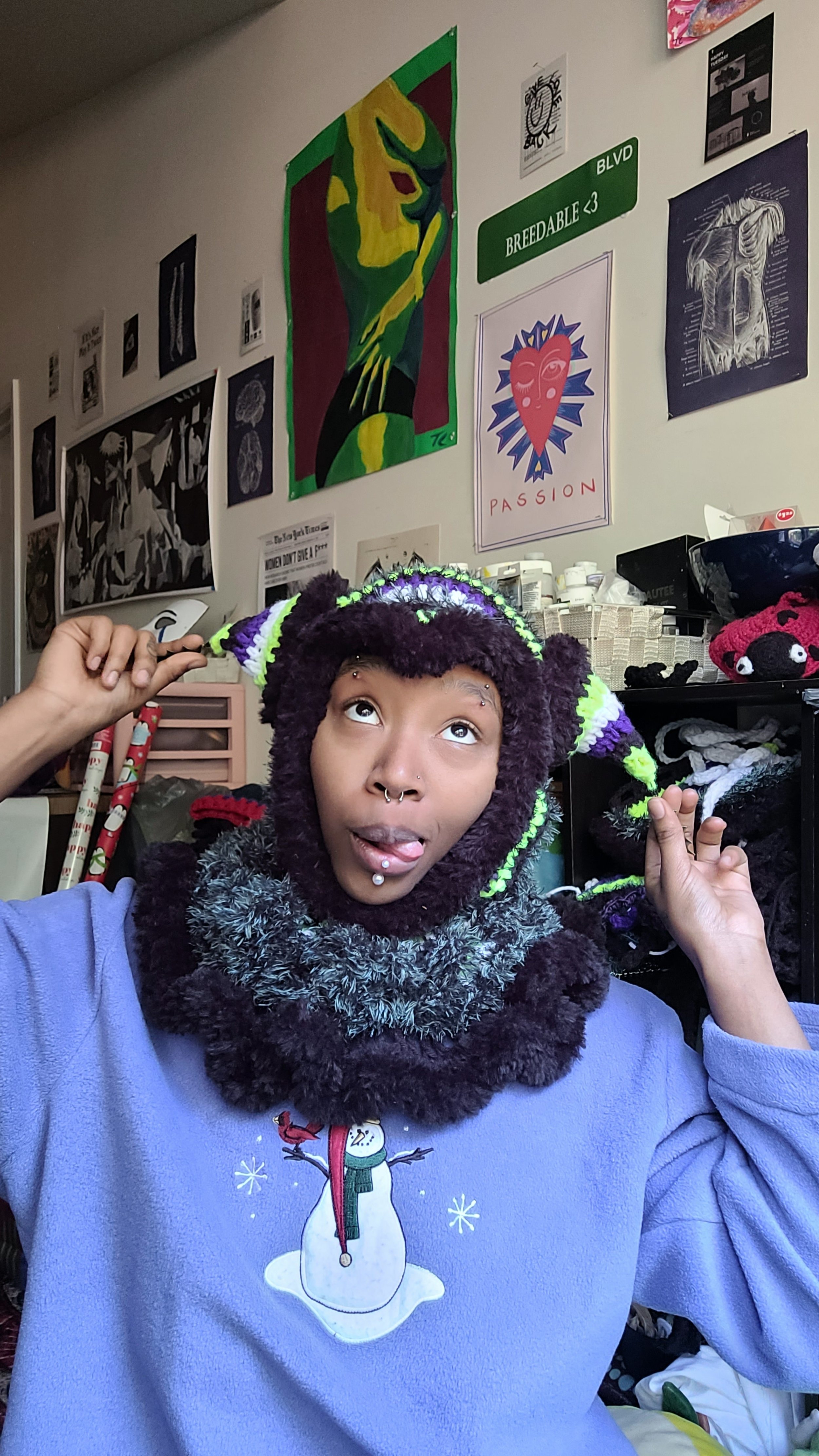 A person with purple and green jester hat, fuzzy gray scarf, and pierced nose and lip, making a playful face with tongue out and eyes looking upward in a room decorated with various artwork and posters on the wall.