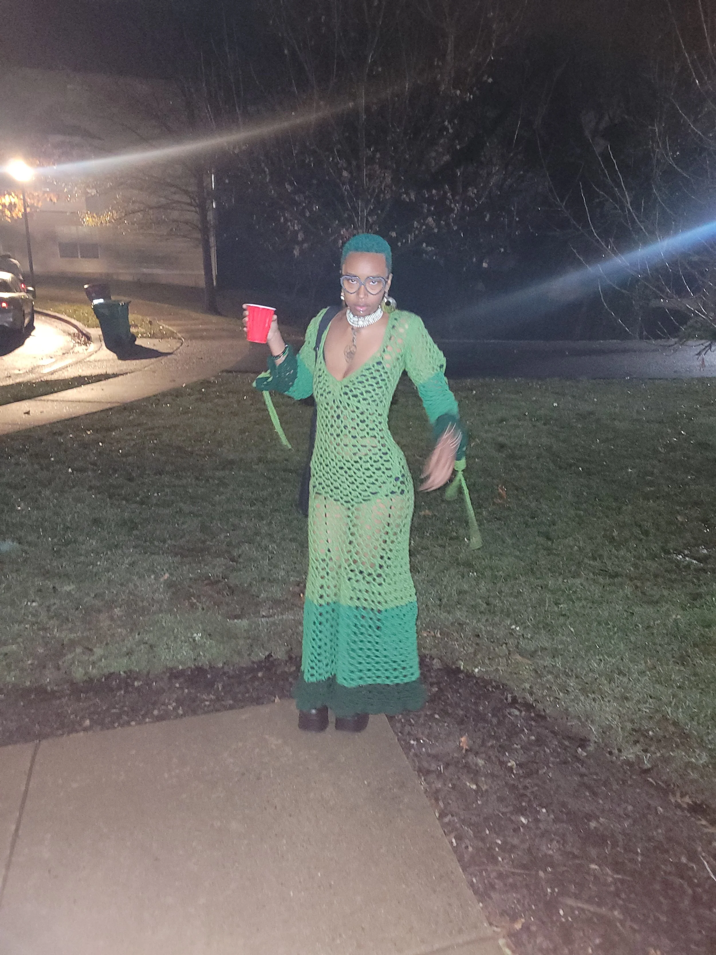 A person with short hair, wearing glasses, a green crocheted dress, and jewelry, standing outside at night, holding a red cup.