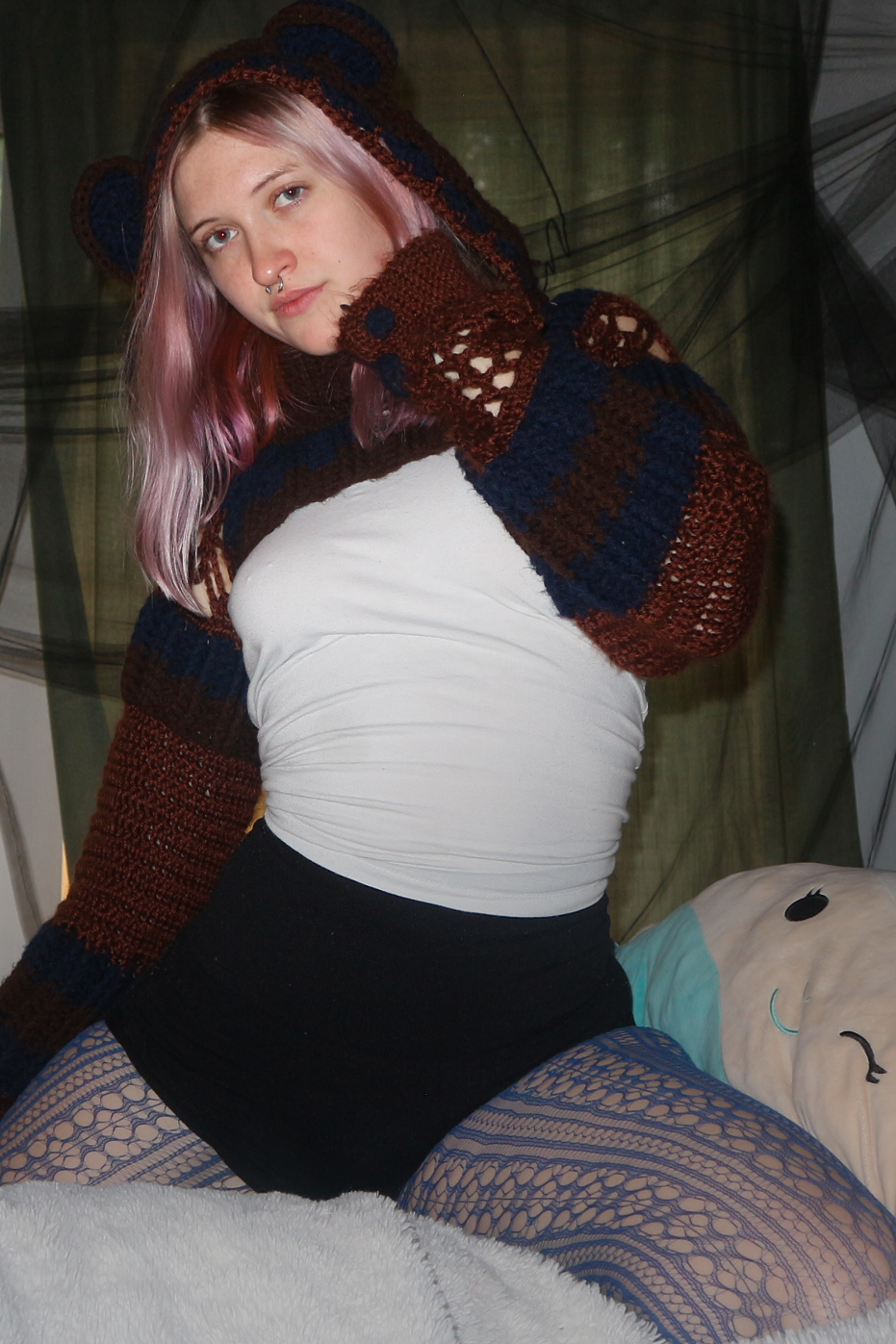 A young woman with pink and blonde hair posing indoors. She is wearing a white top, black shorts, patterned fishnet tights, and a colorful crocheted hoodie. She is sitting on a bed with a plush pillow and black netting in the background.