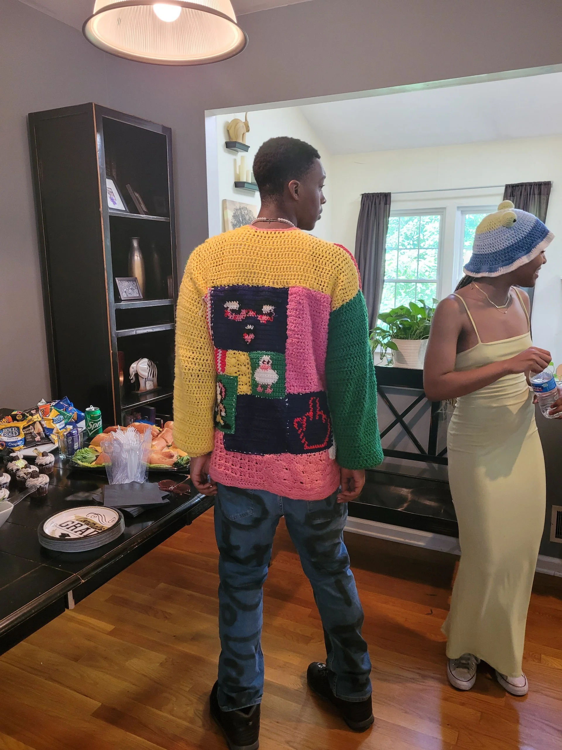 A person facing away from the camera wearing a colorful crochet sweater with patchwork designs, including a face, a peace sign, and a middle finger. They are standing in a room with two women, one wearing a light yellow dress and a knitted hat, and t