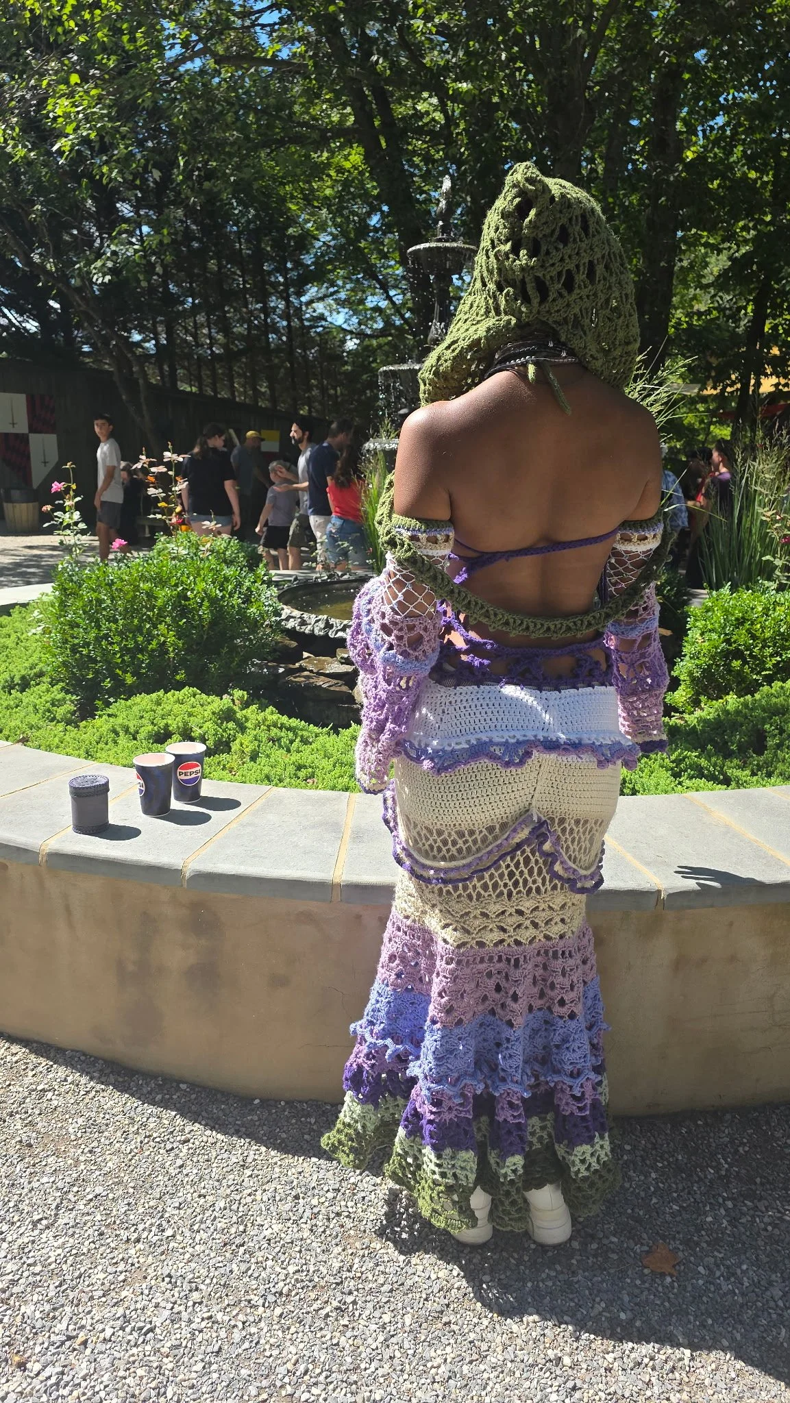 Person wearing colorful crochet dress and green knit head covering standing outdoors near a garden fountain, with people in line or walking in the background.