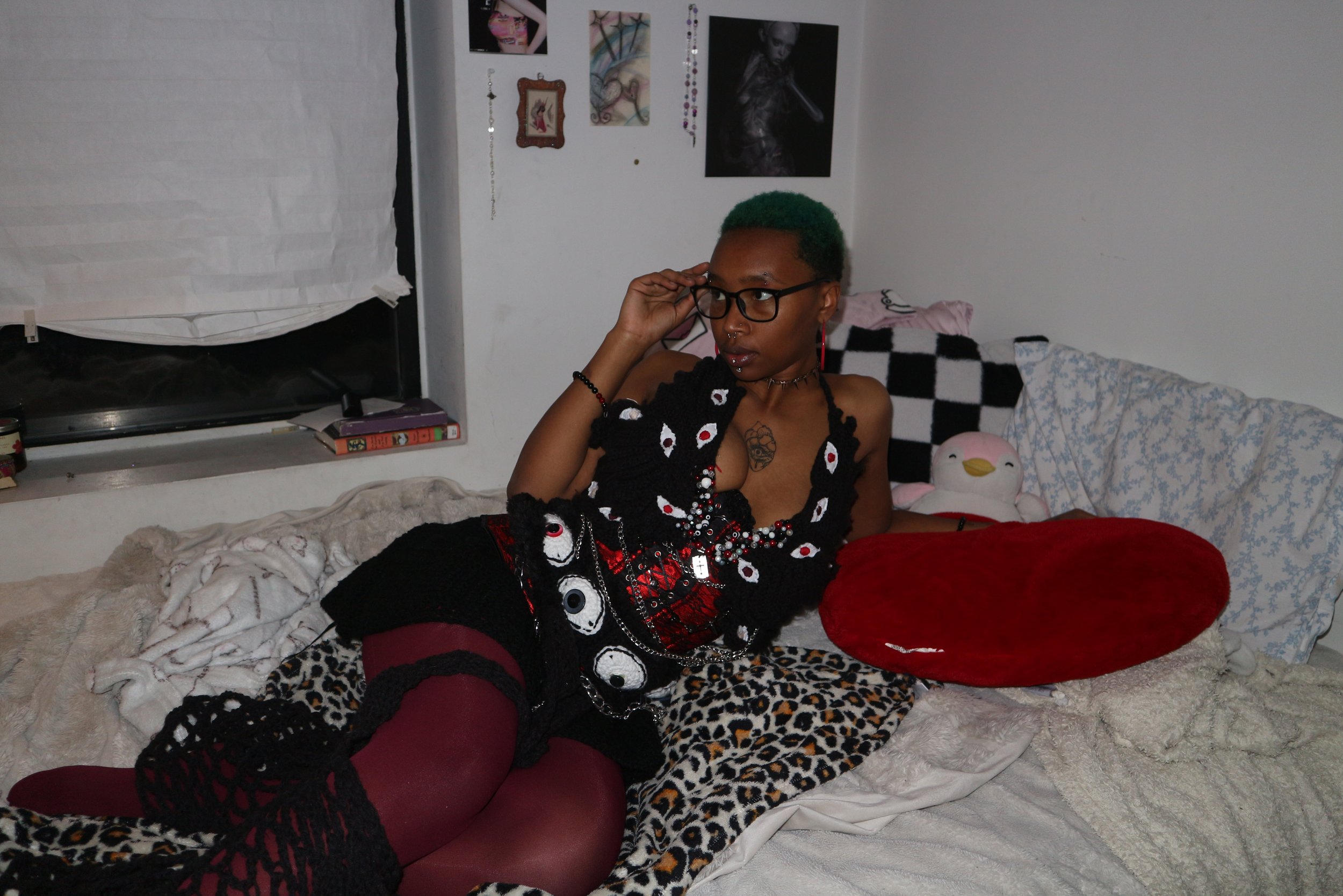 A person with green hair, glasses, and multiple piercings, lying on a bed with leopard print sheets, a red heart-shaped pillow, and various stuffed animals, in a room decorated with artwork and hanging jewelry.