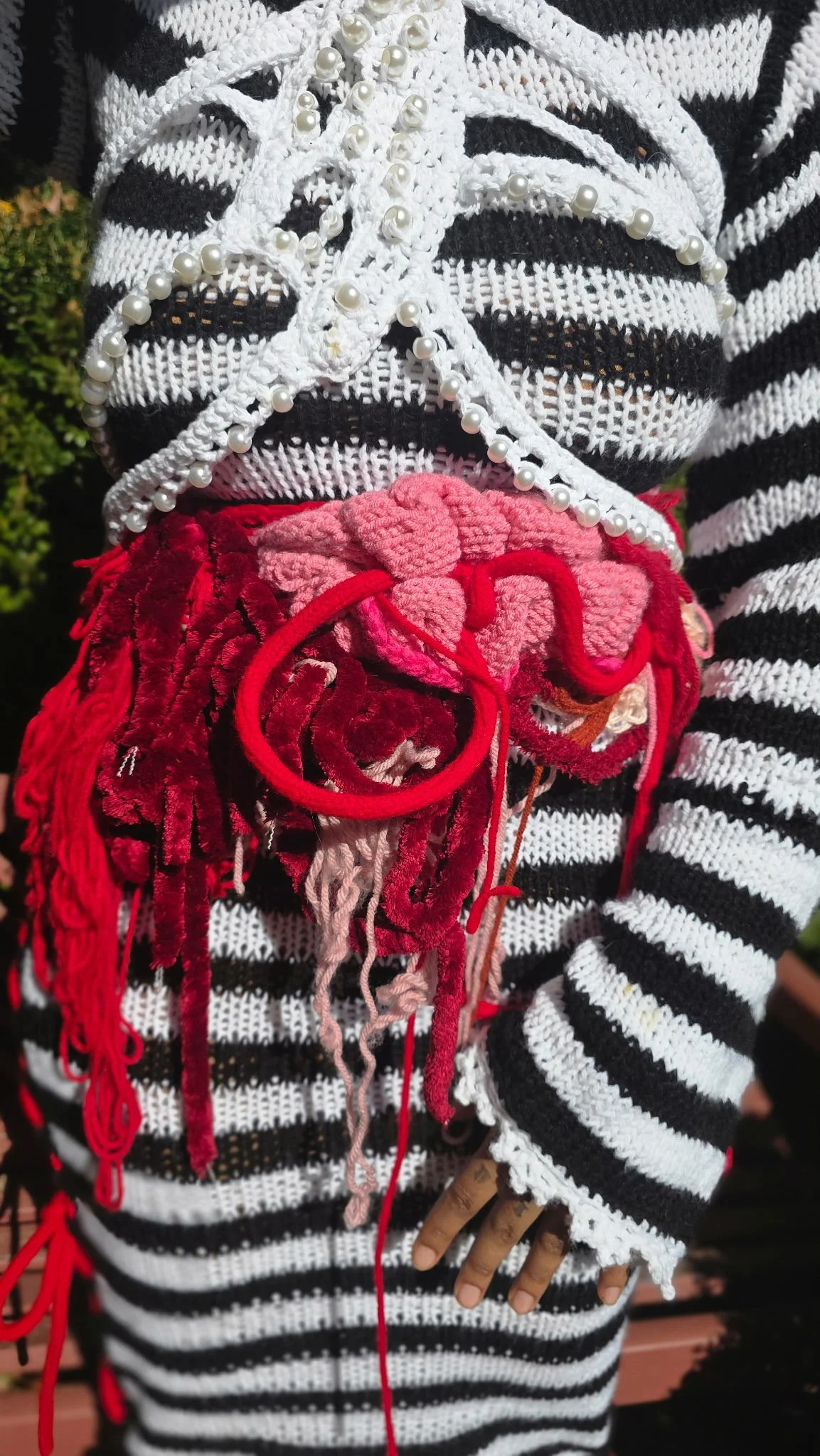 Close-up of a person's torso in a black and white striped knit sweater with pearl embellishments, and a pink fluffy belt with red and pink yarn decorations, with a background of green foliage.