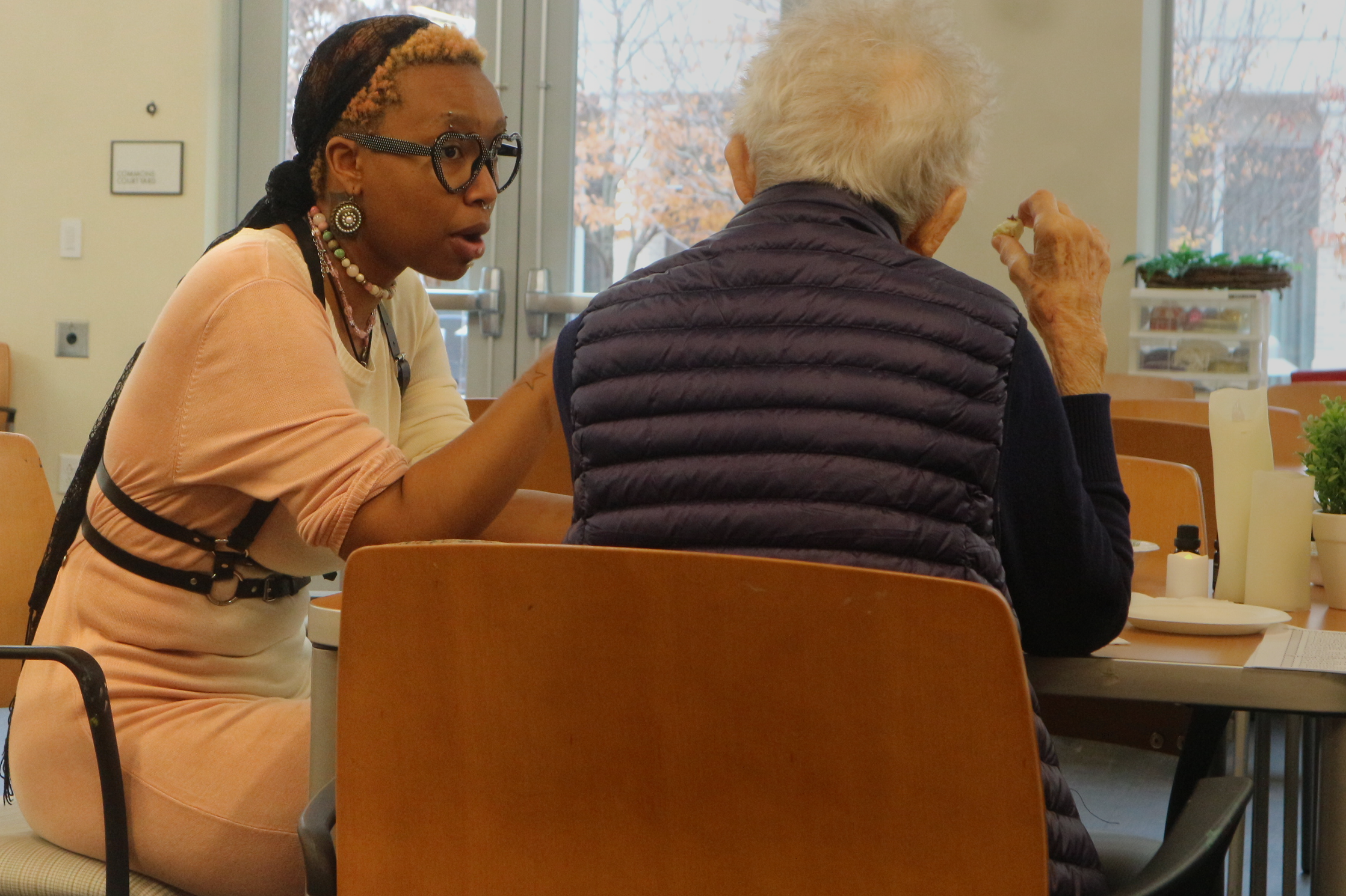 A young woman with glasses, jewelry, and braided hair helps an elderly woman with white hair, sitting at a table in a bright room with large windows, potted plants, and a door with glass panels.
