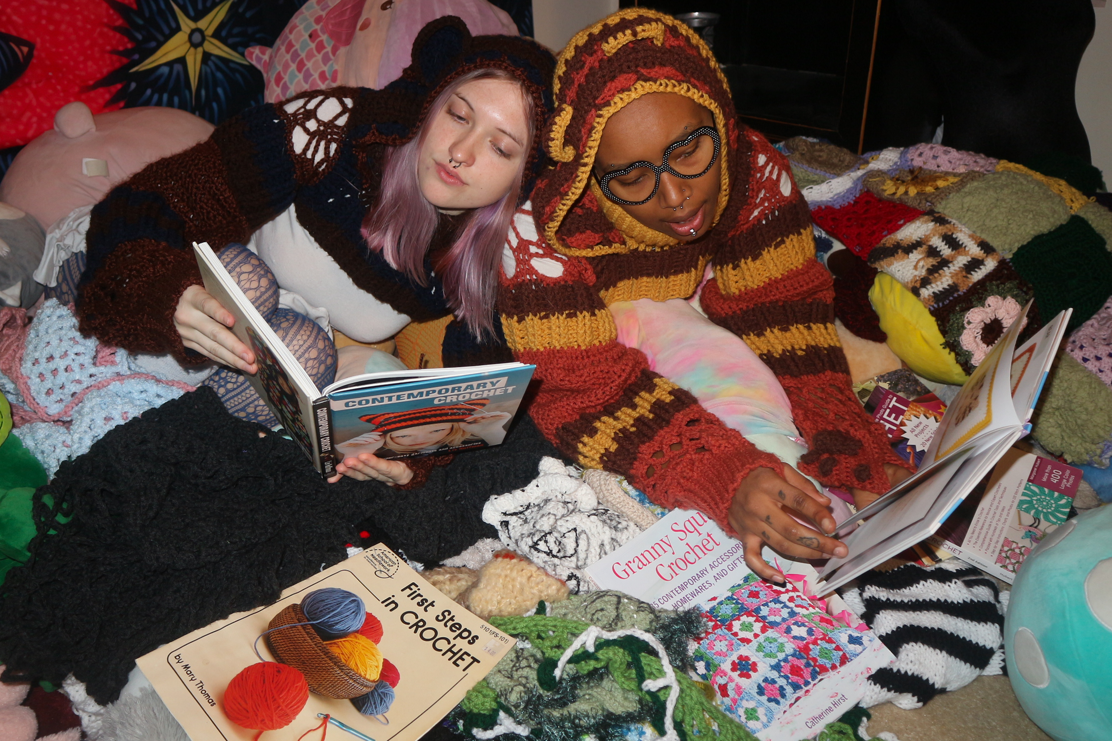 Two women, one with purple hair and glasses, and the other with a knit hoodie, are lying on a pile of colorful crocheted blankets and pillows, reading crochet books amidst homemade crocheted items and yarn.