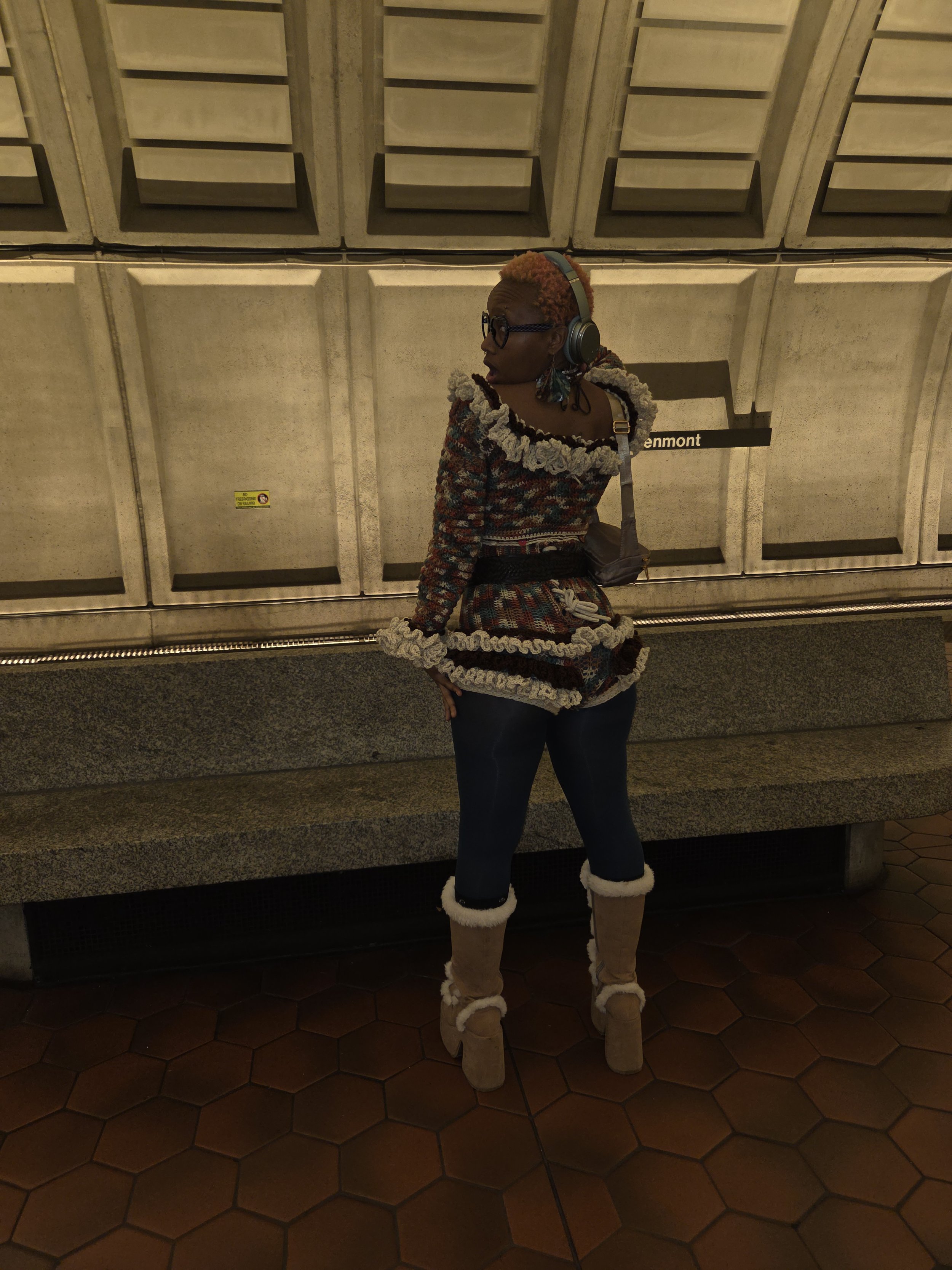 A woman with short, colorful hair wearing glasses, headphones, a multicolored knitted sweater with ruffled edges, dark jeans, and beige boots standing at a subway station.