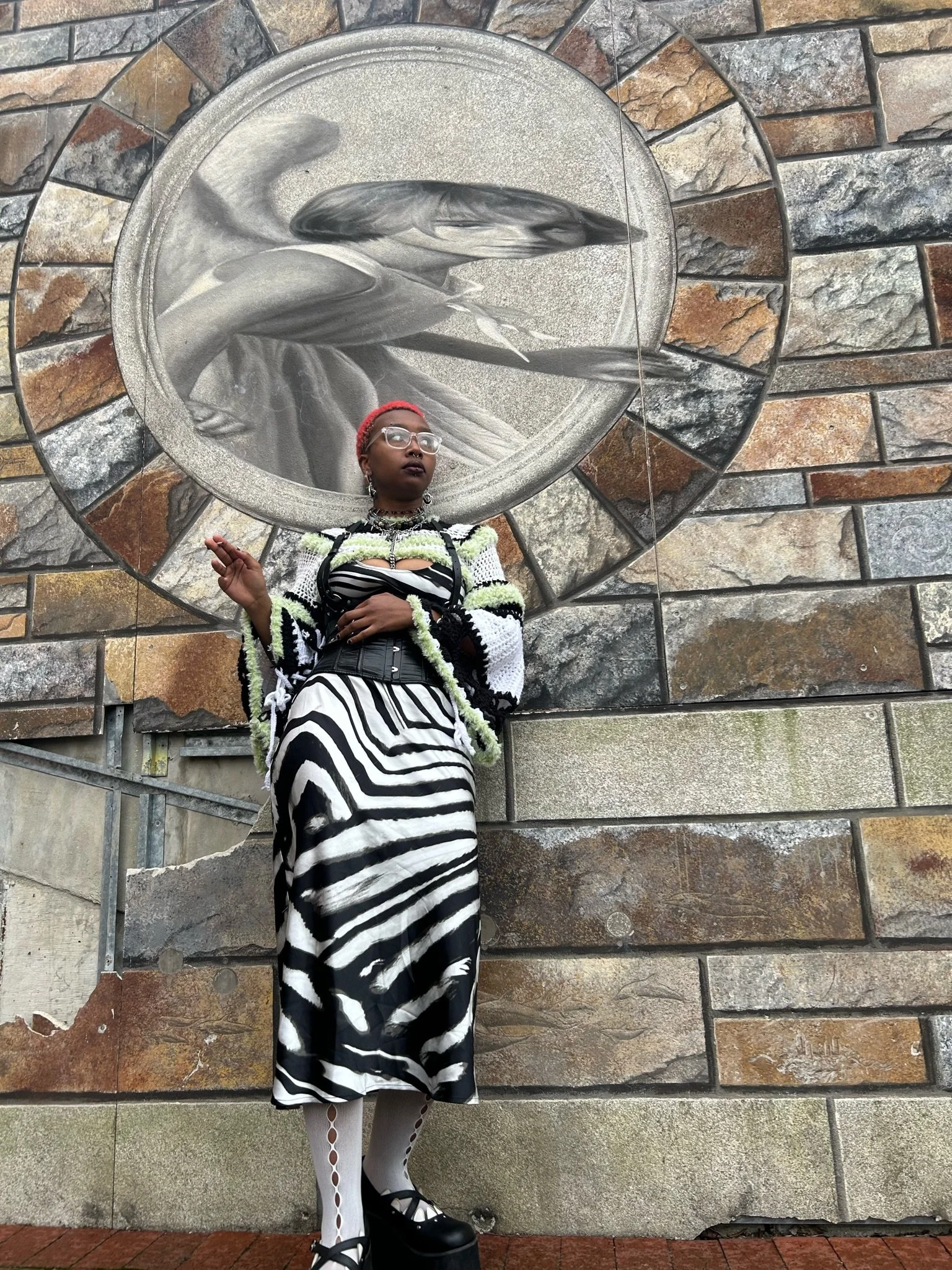 A person standing in front of a large wall mural of a bird, wearing a black and white zebra-striped skirt, white tights with cut-out circles, black platform shoes, glasses, and a colorful knitted sweater.
