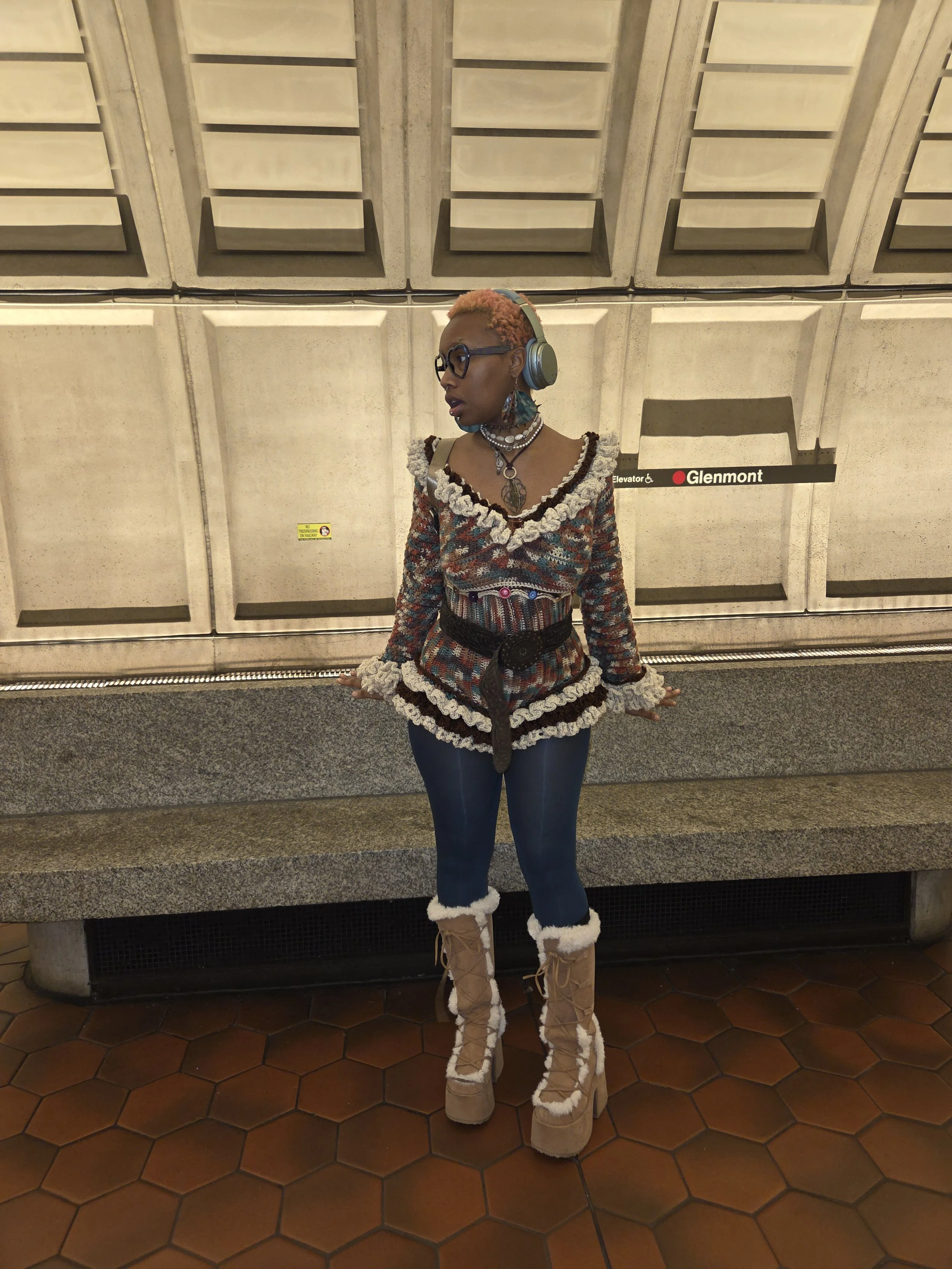 A woman standing in an underground transit station, wearing headphones, glasses, a colorful textured top with lace embellishments, jeans, and high-heeled boots lined with fluffy material, with a background of concrete walls and tiled floor.