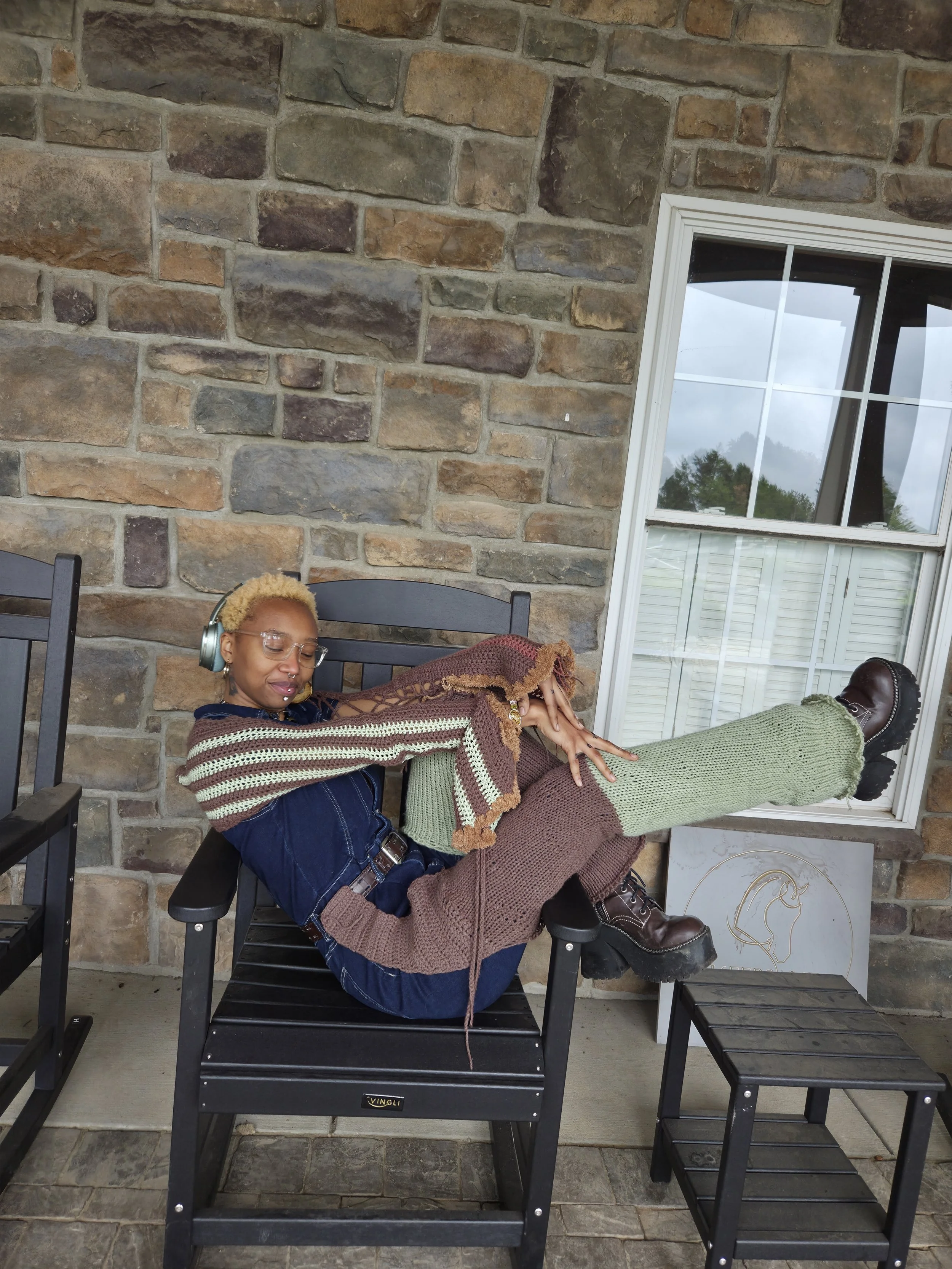 Person sitting on a black chair with their feet up on a small black table, wearing colorful striped and knitted clothing, listening to headphones, with a stone wall and window behind them.