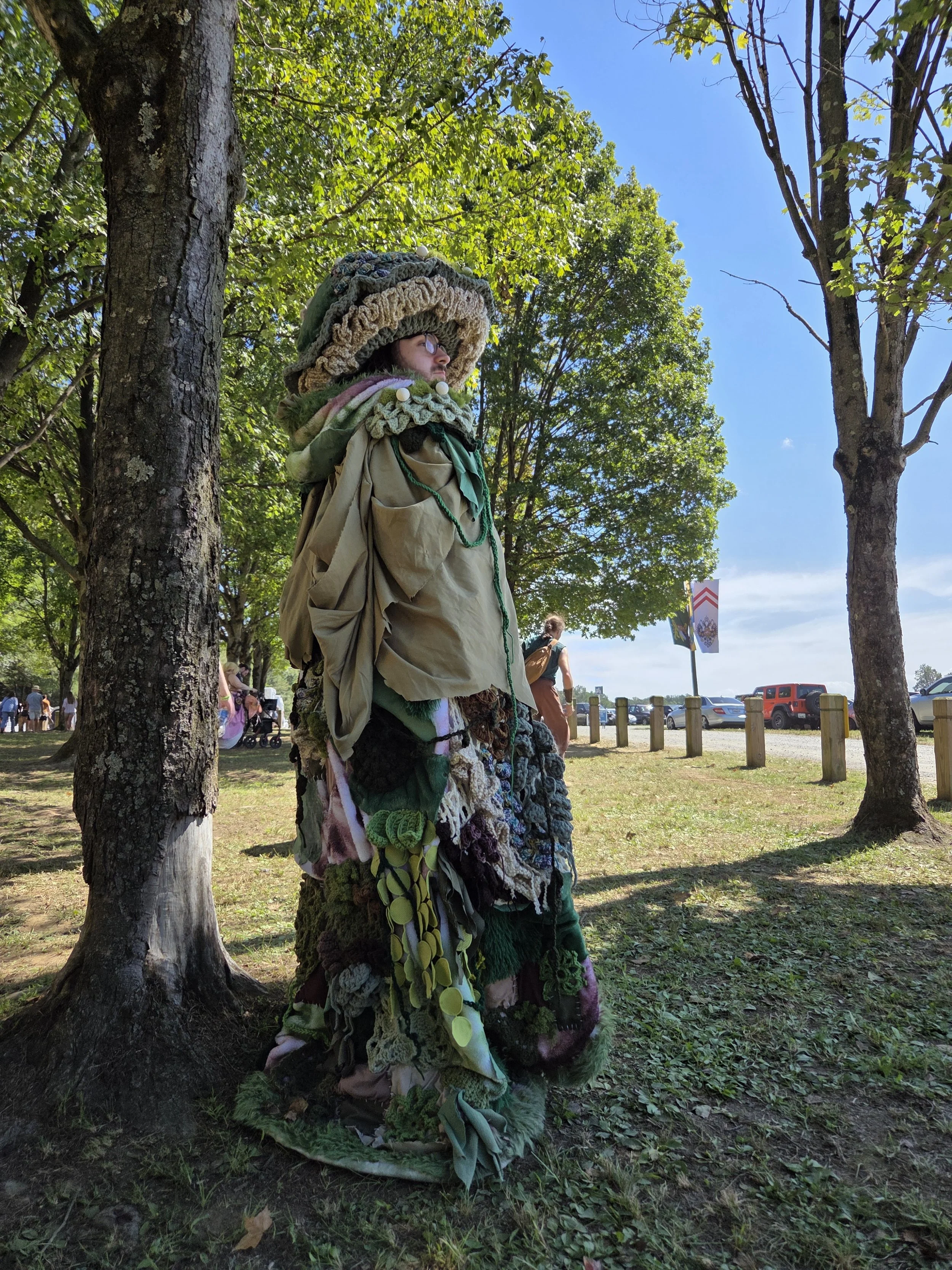 Person dressed in a costume made of various textiles, yarns, and fabrics, standing outdoors between trees near a parking lot on a sunny day.