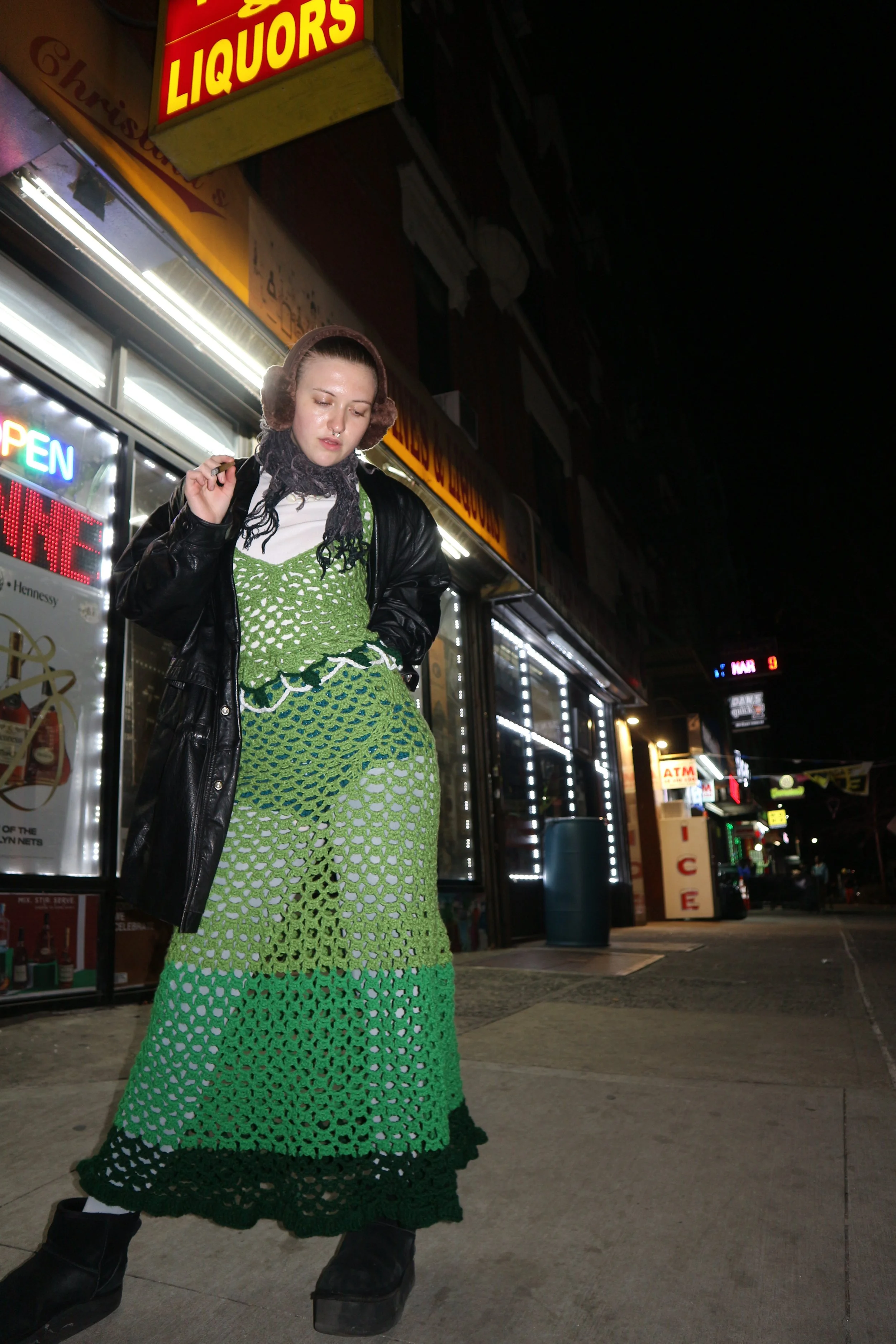 A woman wearing a black leather jacket and a green crochet dress standing on a city sidewalk at night in front of a liquor store with bright neon signs, including an 'OPEN' sign.