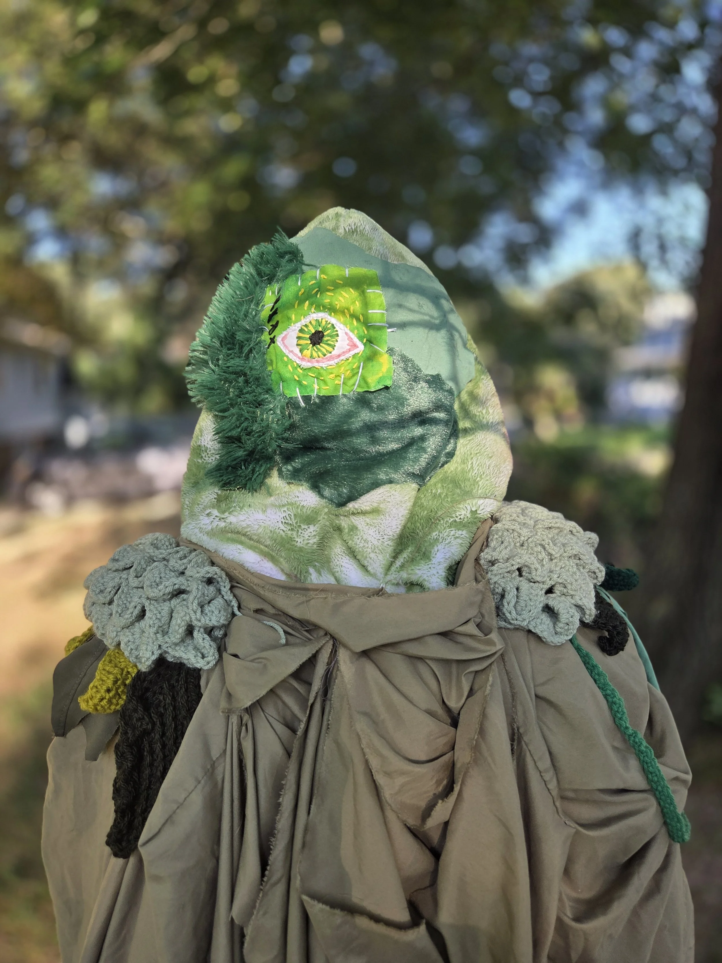 A scarecrow with a patchwork face featuring a large green eye, dressed in a brown jacket, with crocheted fabric details on the shoulders, standing outdoors with a blurry background of trees and blue sky.