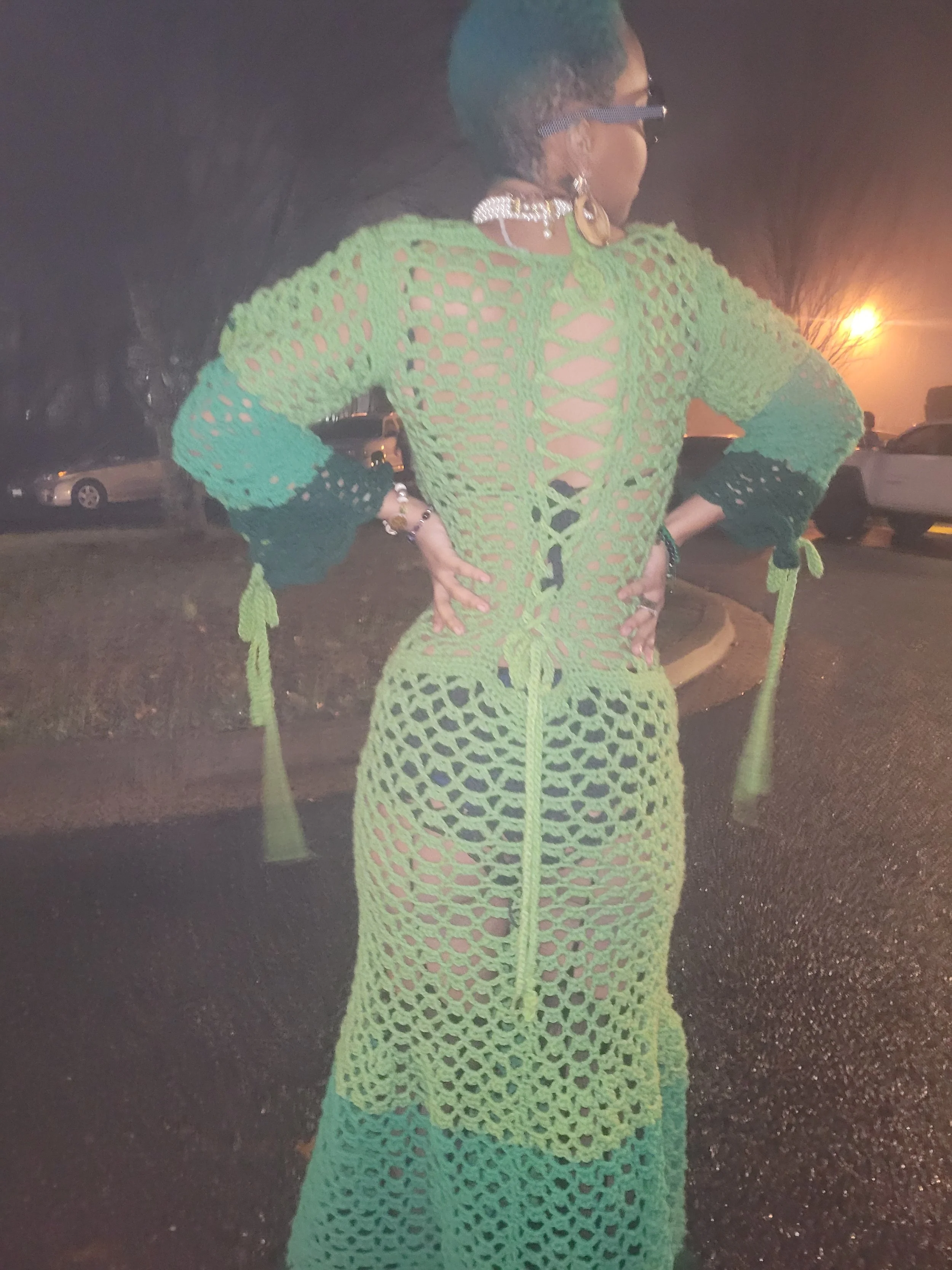 A woman wearing a brightly colored crocheted dress with cutout designs, standing outdoors at night with her hands on her hips.