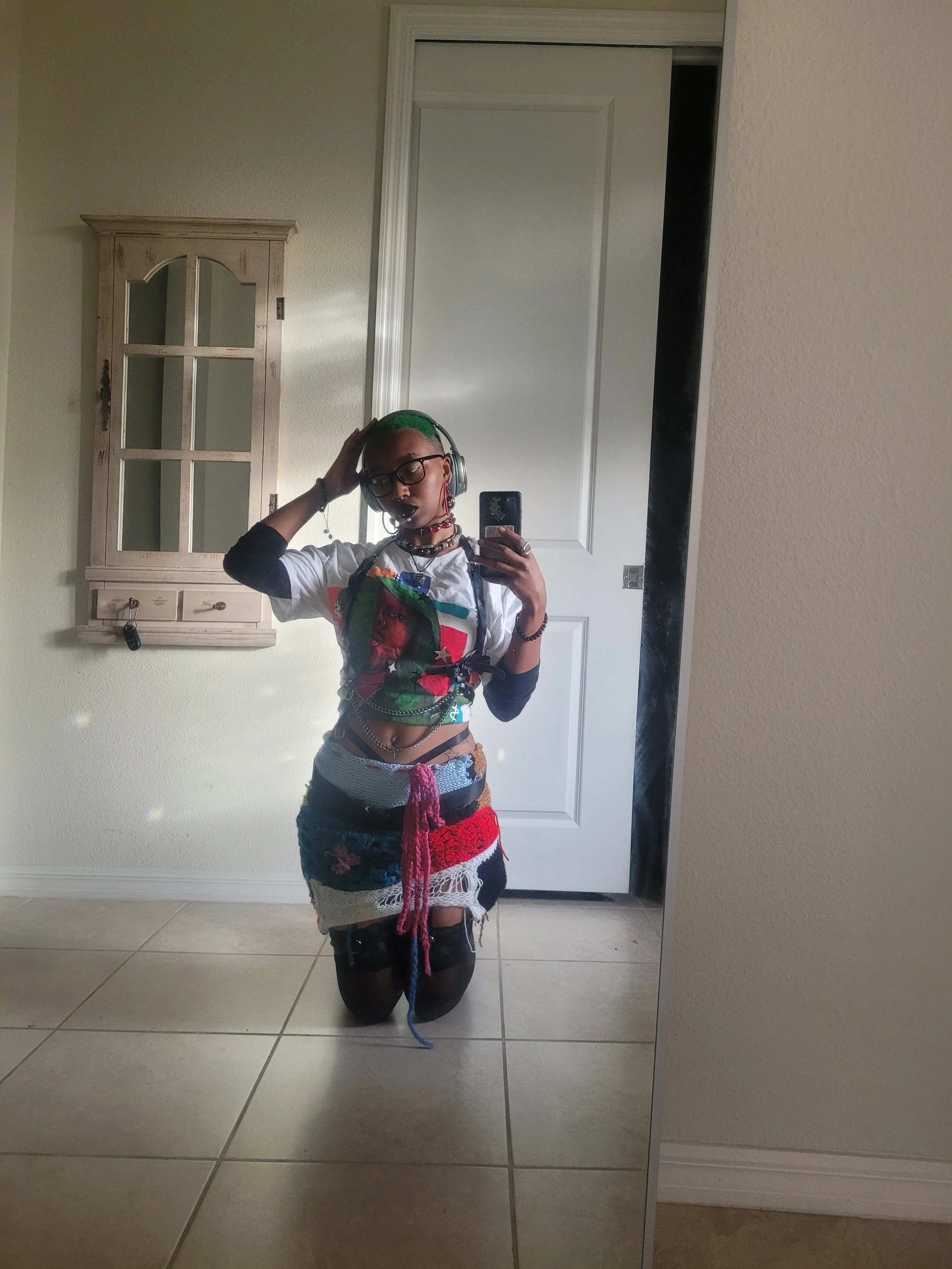 Person with green hair taking a mirror selfie inside a room, kneeling on the floor, wearing headphones, glasses, a t-shirt, and a colorful, patchwork skirt with various textures and colors.