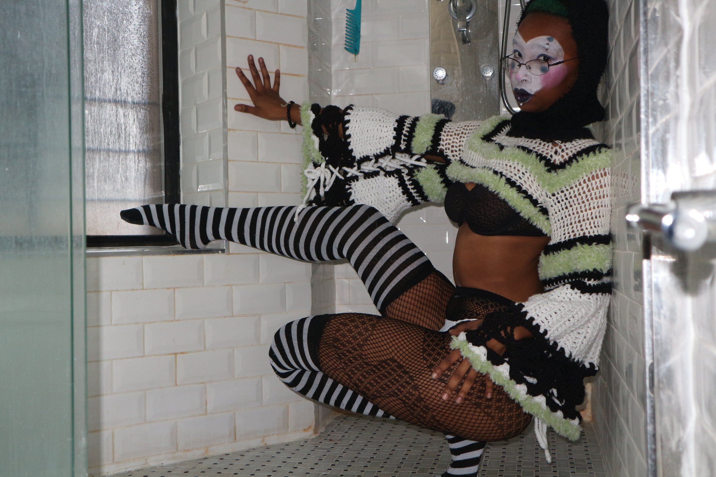 A person wearing black and white striped tights, fishnet stockings, and a colorful, crocheted sweater with neon green, black, and white stripes, sitting on a bathroom or shower floor with white tiled walls. They have makeup on their face resembling a clown or mime, with glasses and dark lipstick, and are posing for a photo.