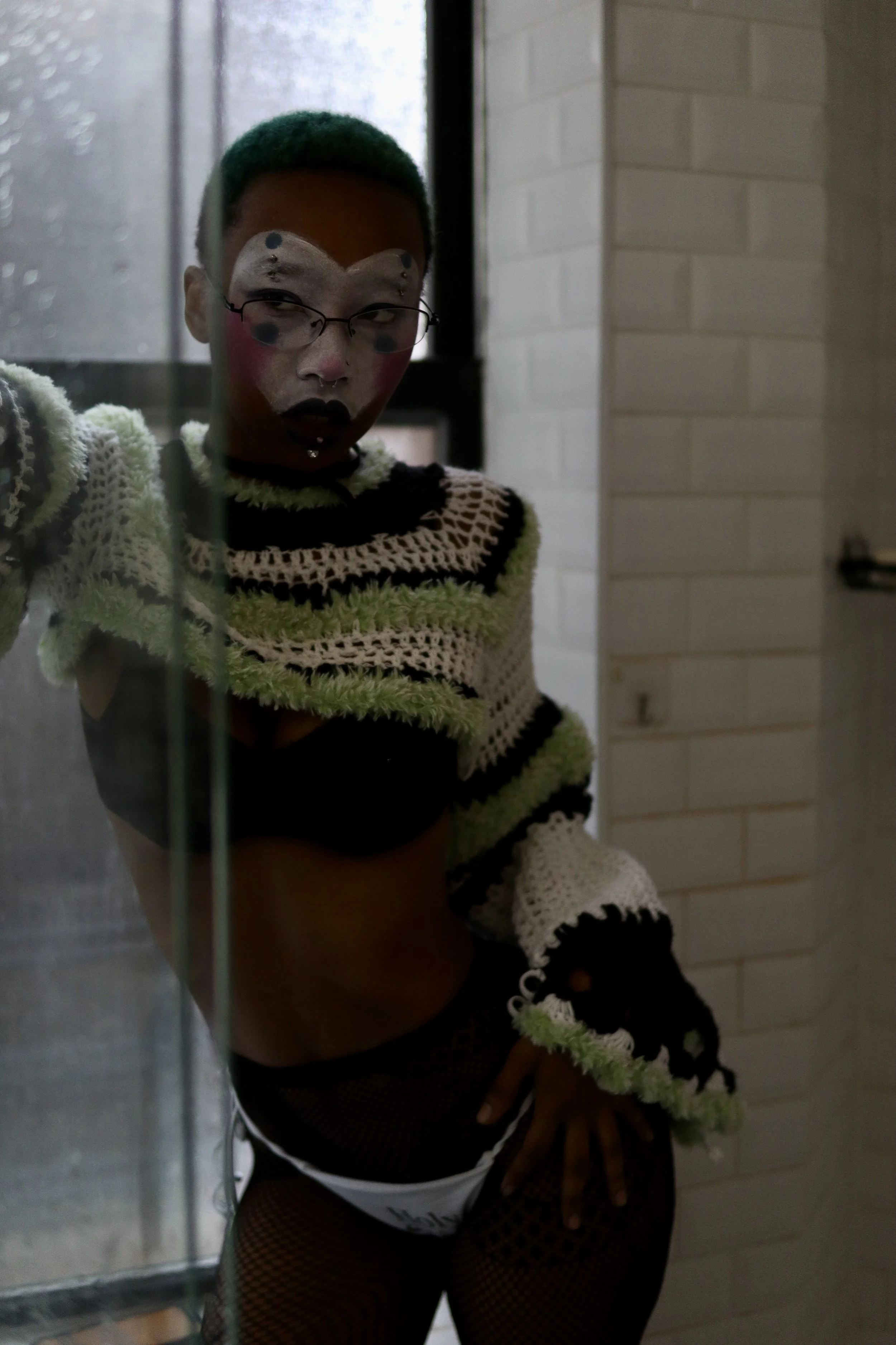 Person with short, colored hair and face paint, wearing a crochet sweater, fishnet tights, and underwear, standing in a dimly lit room near a window and a brick wall.