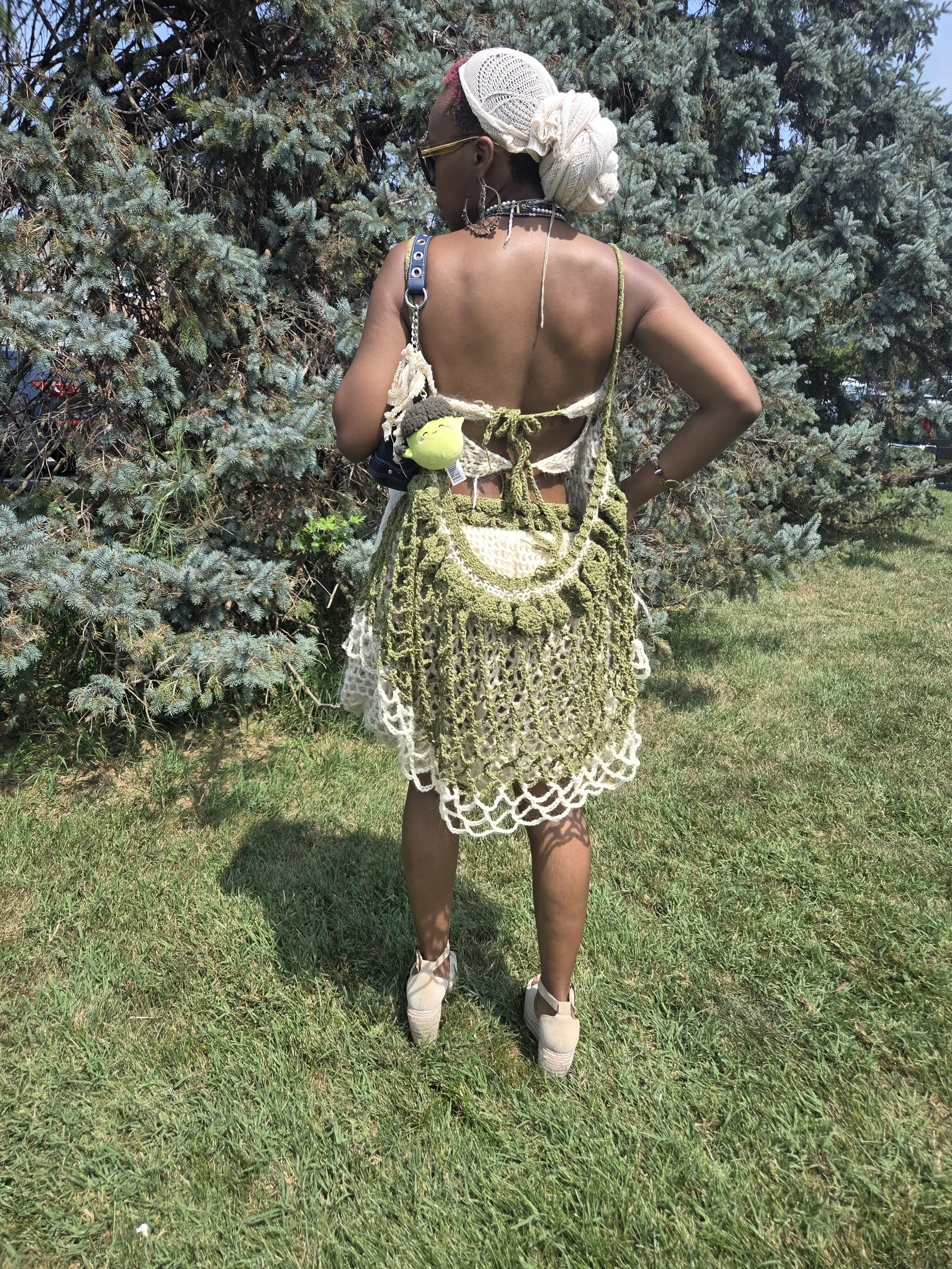 A woman standing outdoors on grass with evergreen trees in the background. She is wearing beige wedge shoes, sunglasses, large hoop earrings, and a lace headscarf. She has on a backless top and carries a green, handmade, crocheted bag.