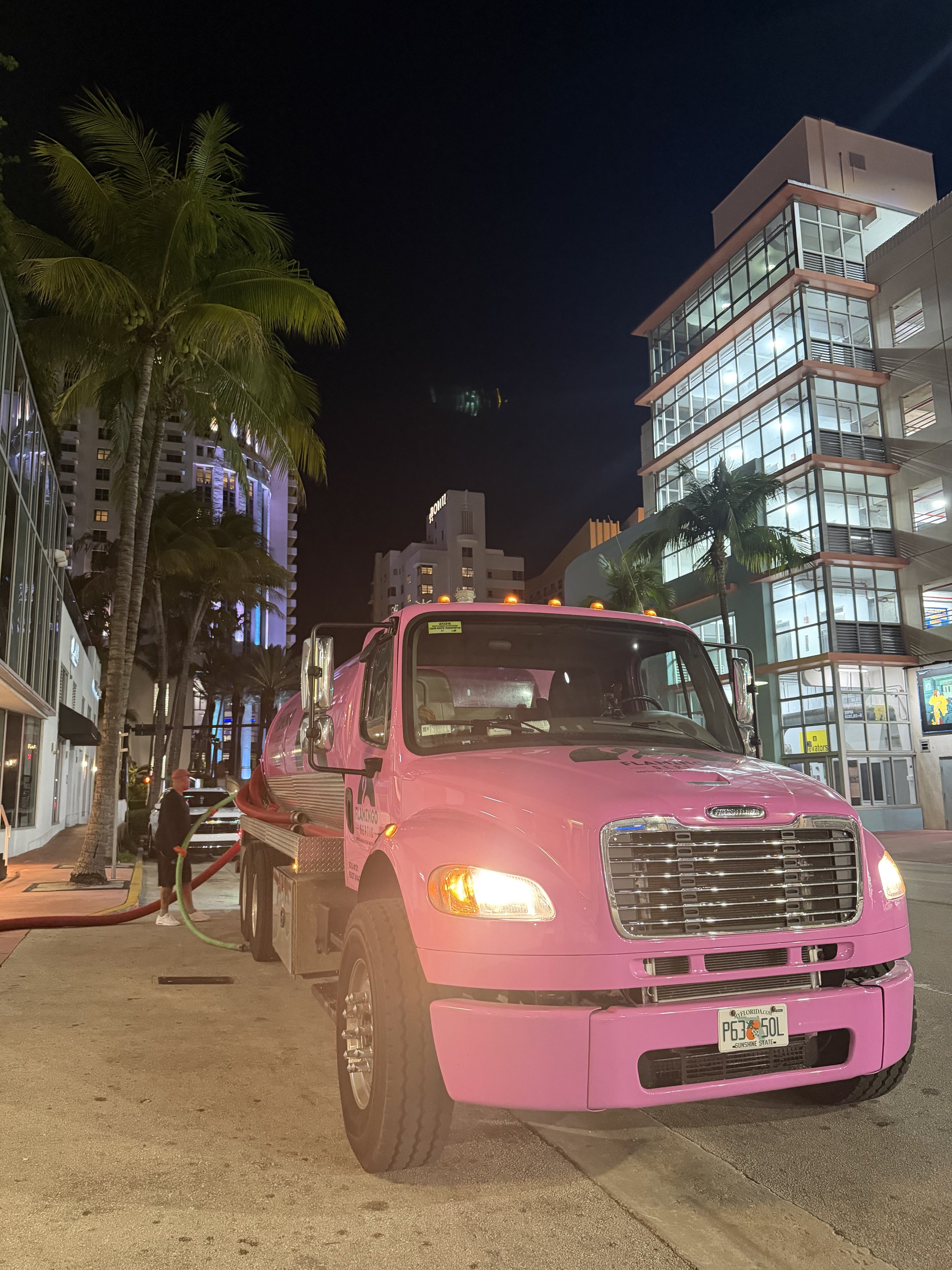 A pink grease trap truck parked on a city street at night with palm trees and modern buildings illuminated in the background.