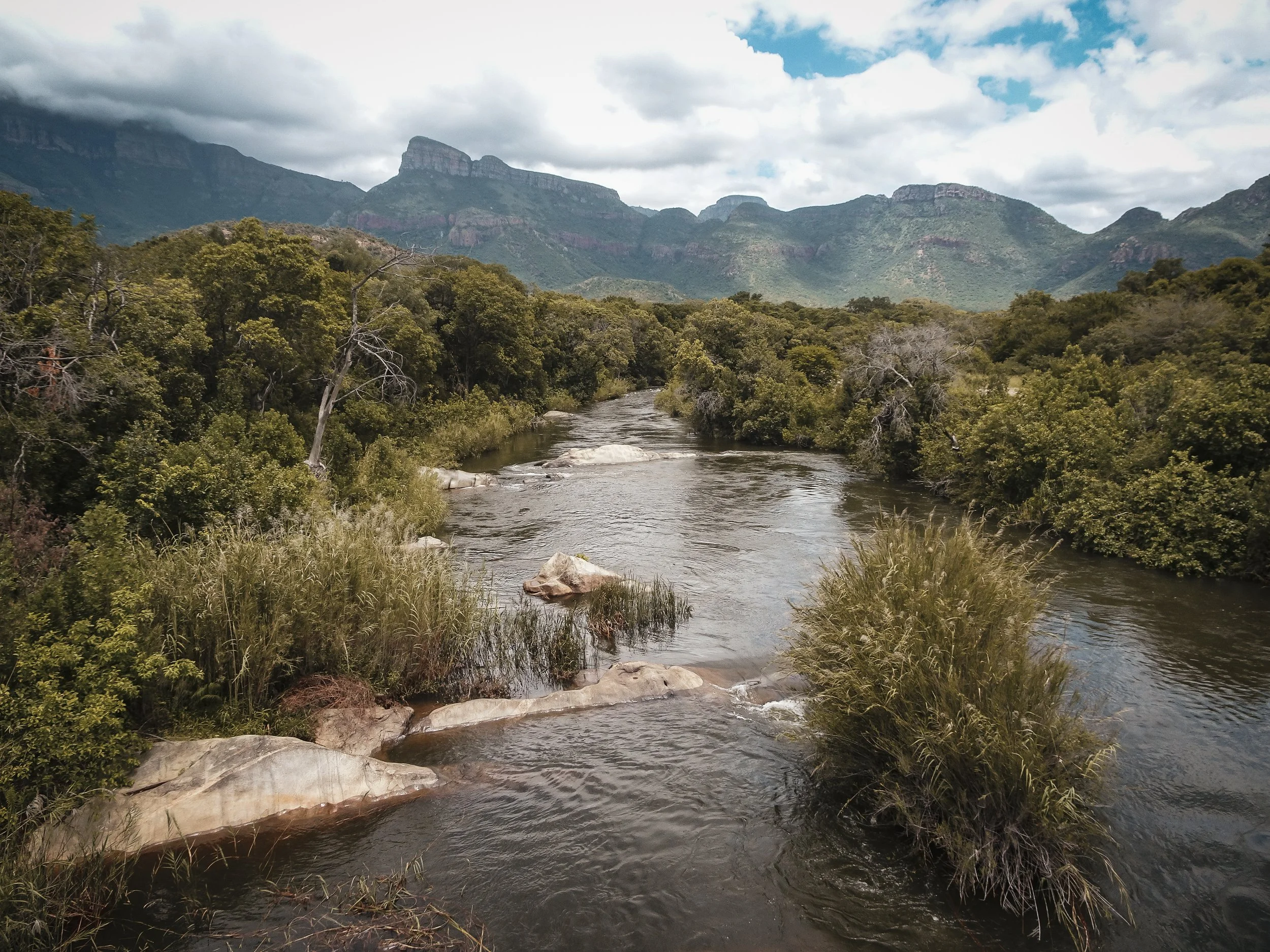 A river flowing through a lush green landscape with mountains in the background under a cloudy sky.