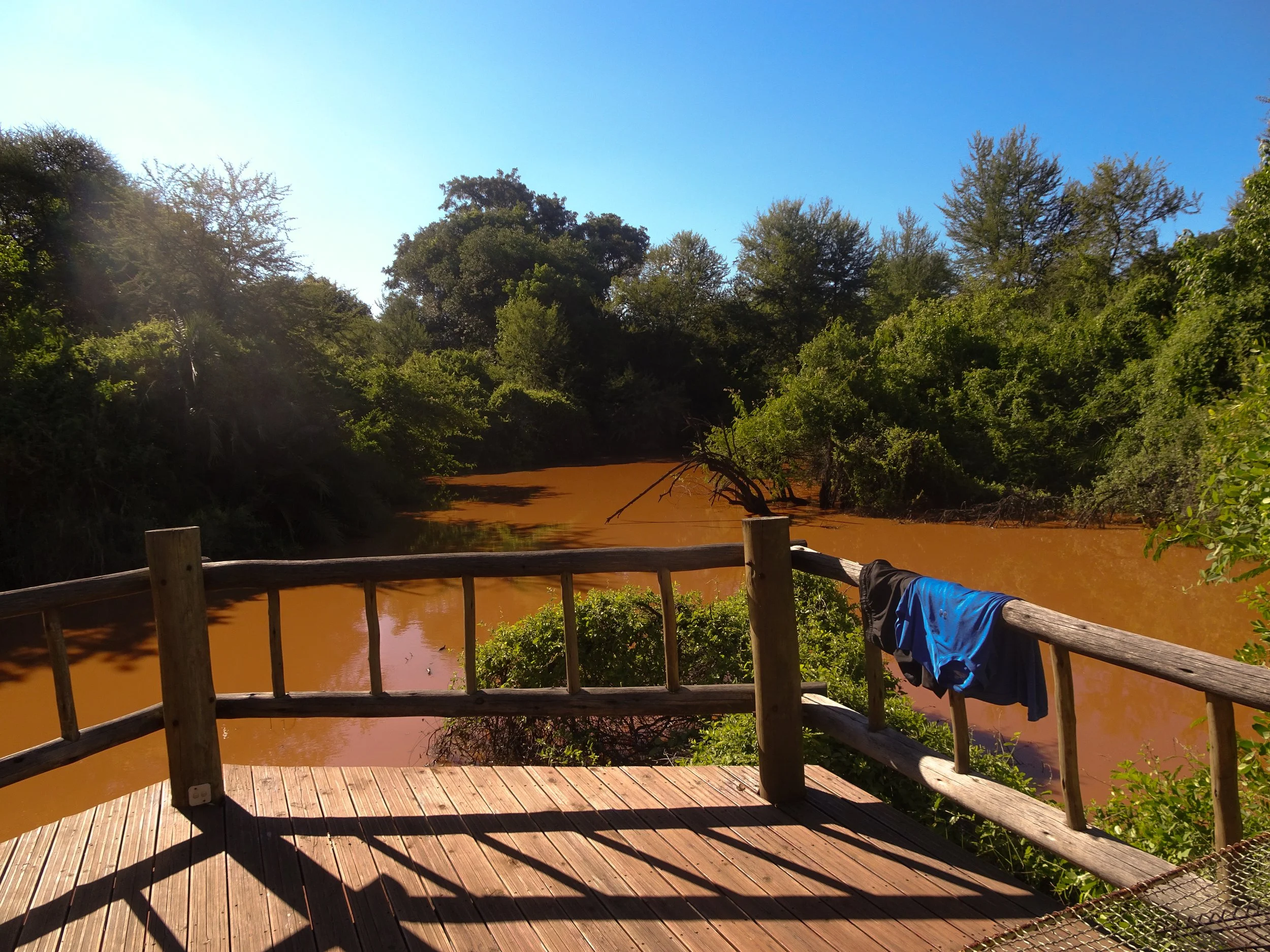 A wooden deck overlooking a river with muddy water, surrounded by lush green trees under a clear blue sky. A blue shirt is draped over the deck railing.