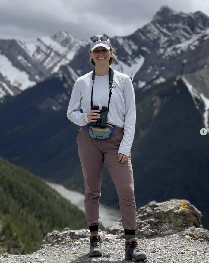 A woman standing on a rocky outcrop with mountain peaks in the background, dressed for hiking with binoculars around her neck and a camera hanging from her neck.