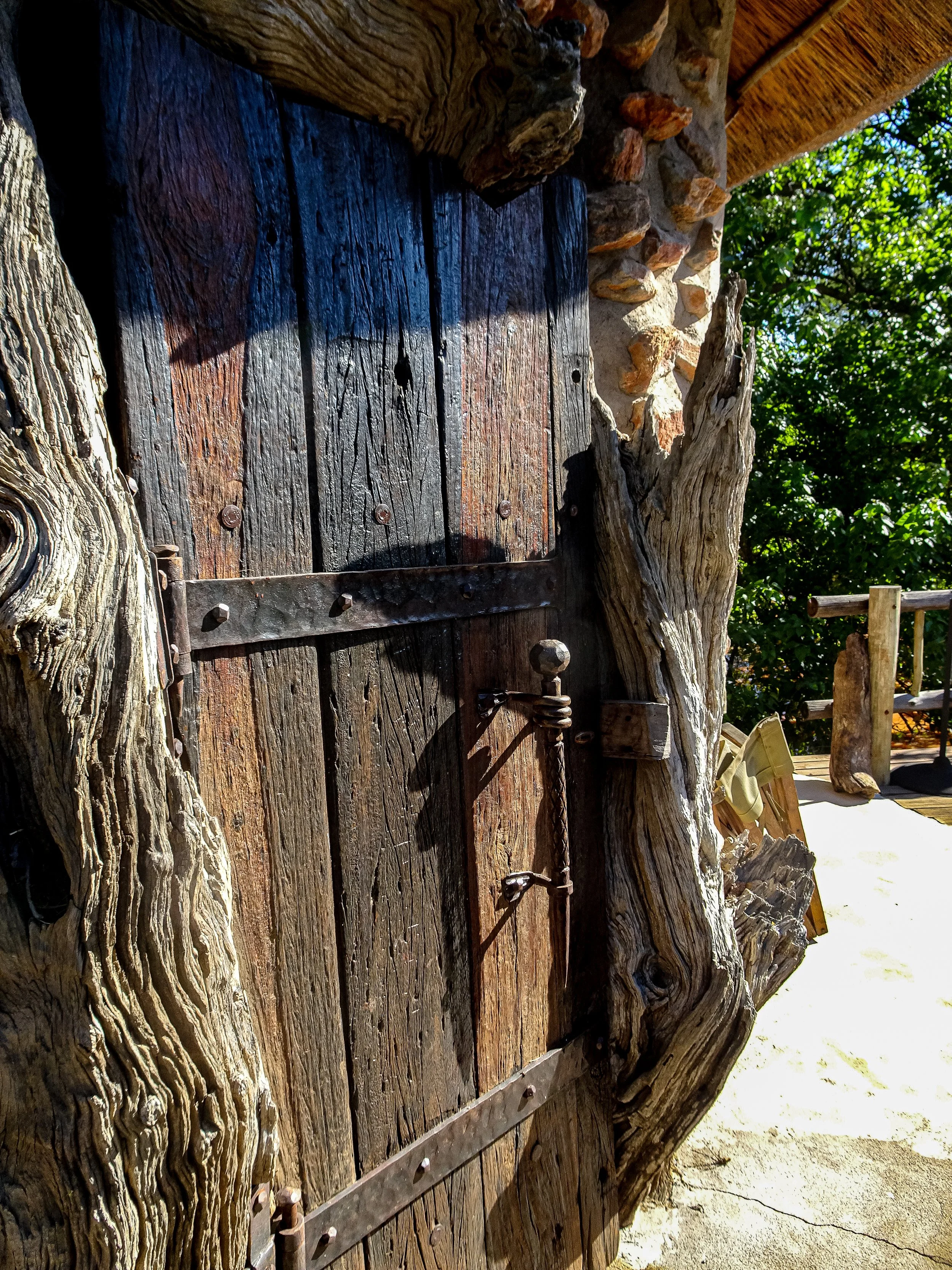 Close-up of a rustic wooden door with iron hinges, surrounded by gnarled wood and stone, outdoors with green foliage in the background.