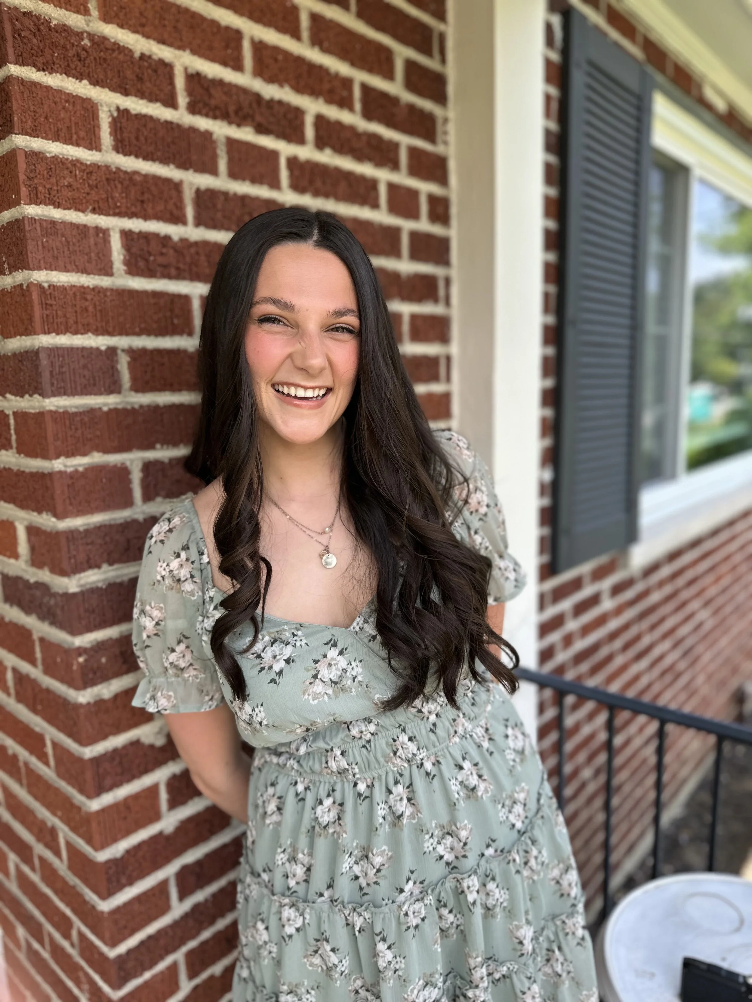 A young woman with long dark hair and a floral dress, smiling and leaning against a brick wall on a porch.