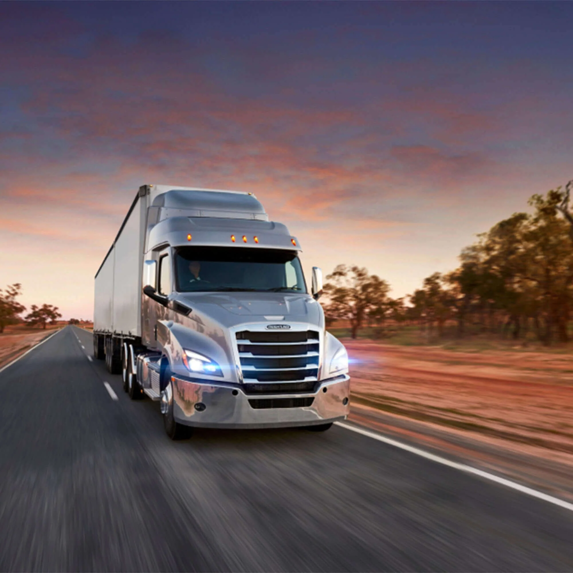 A silver semi-truck driving on an open highway during sunset with trees on the side and a colorful sky.