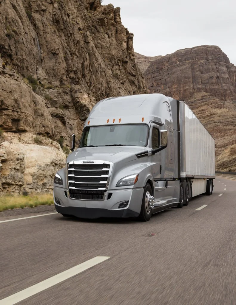 A silver semi-truck driving on a highway through a rocky desert landscape with cliffs and mountains in the background.