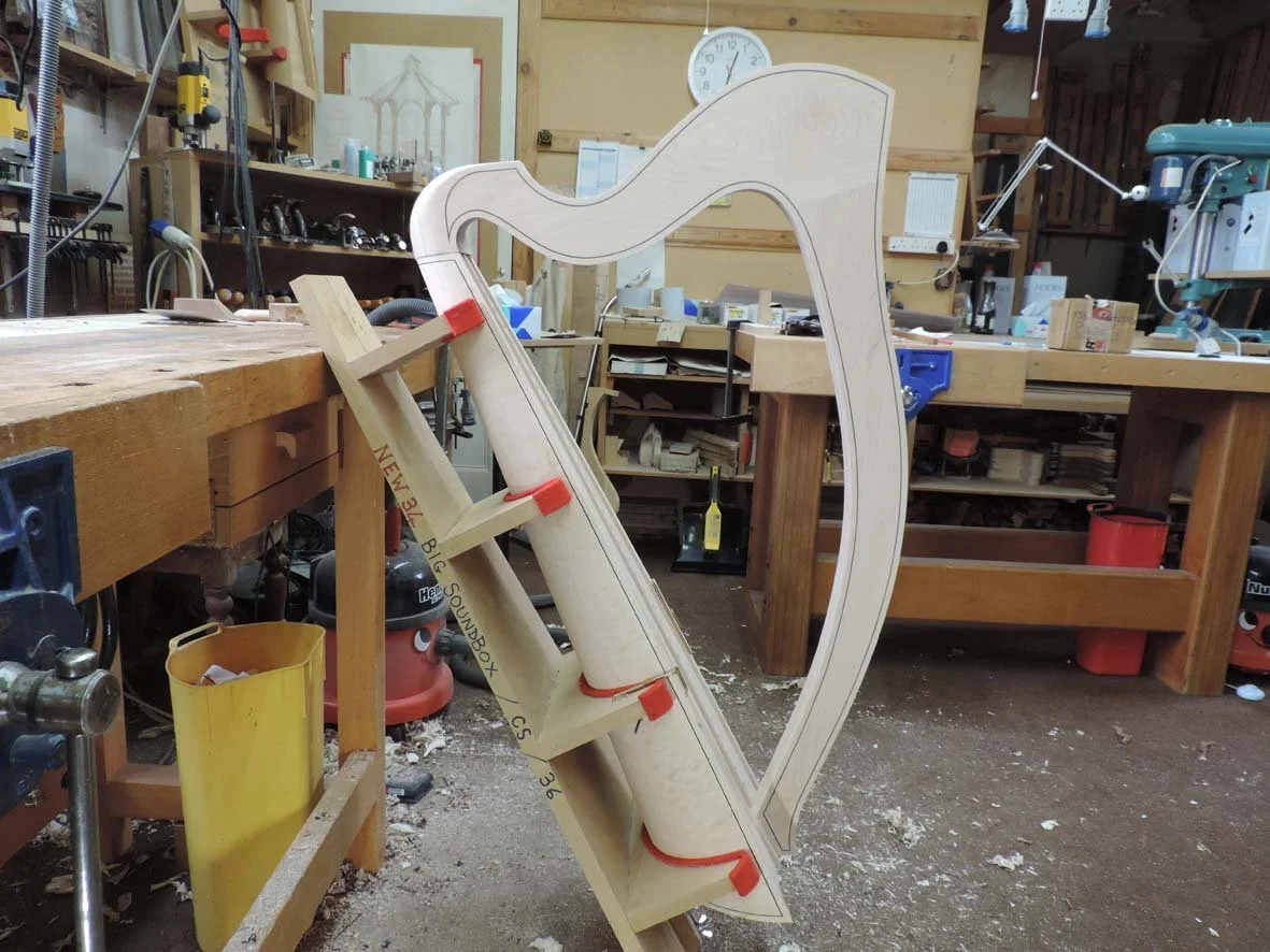 A woodworking workshop with a large harp-shaped piece of wood secured with clamps on a workbench. The workshop is filled with tools, equipment and wooden furniture.
