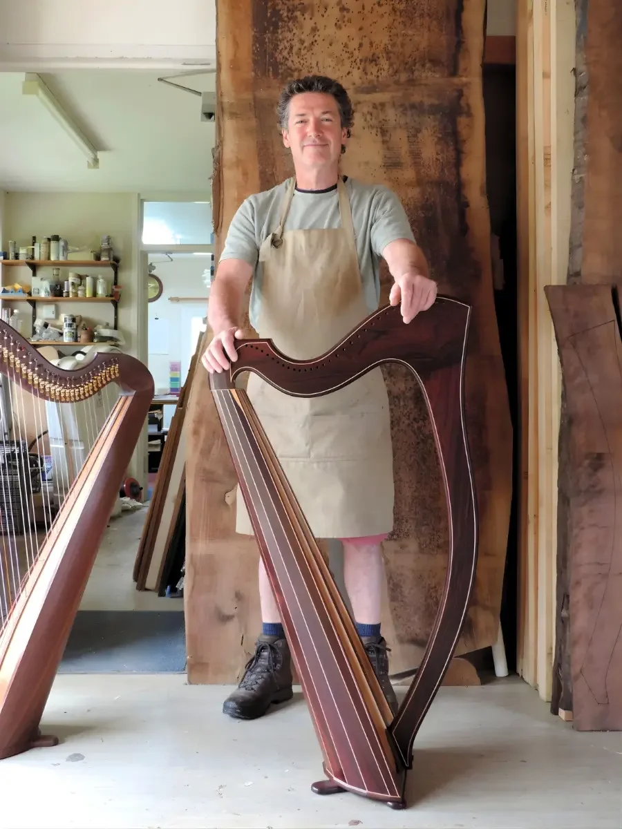 A man standing in a woodworking shop, holding a harp, with another harp partially visible in the foreground. The man is wearing a gray t-shirt, beige apron, and black shoes. The background includes various woodworking tools and supplies.