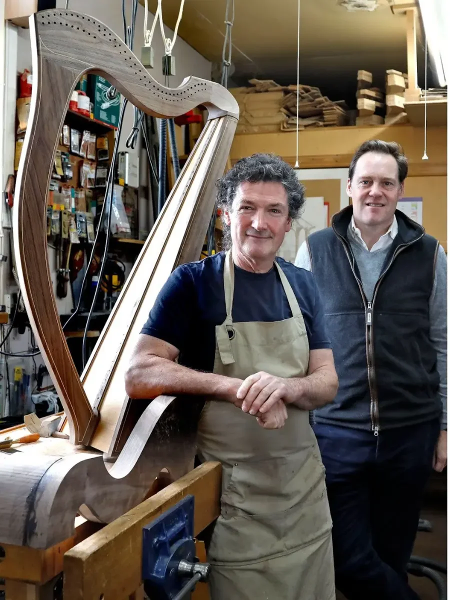 Two men in a woodworking shop with a harp ornament in front of them. One man wears a beige apron, the other a dark waistcoat, and they are surrounded by woodworking tools and materials.