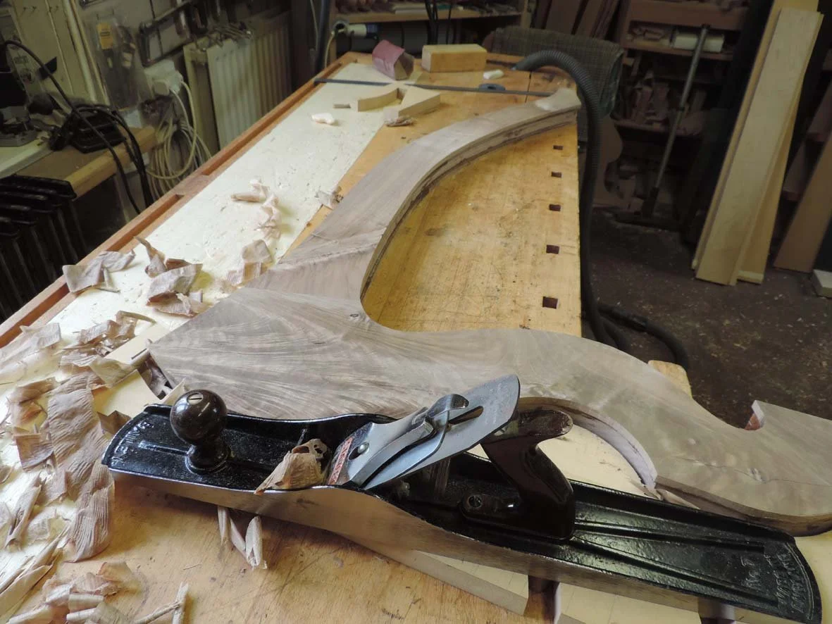 A woodworking workbench with a partially carved large piece of wood, a hand plane, and shavings scattered on the surface in a woodworking shop.