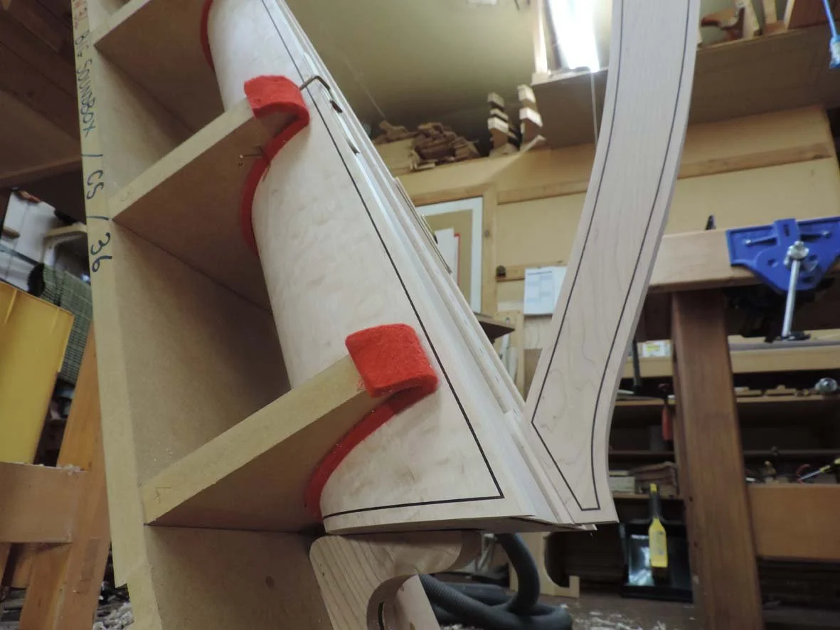 Close-up of a woodworking project in progress, showcasing a curved wooden piece with detailed lines drawn on it, clamped to a workbench with red felt pads for protection.