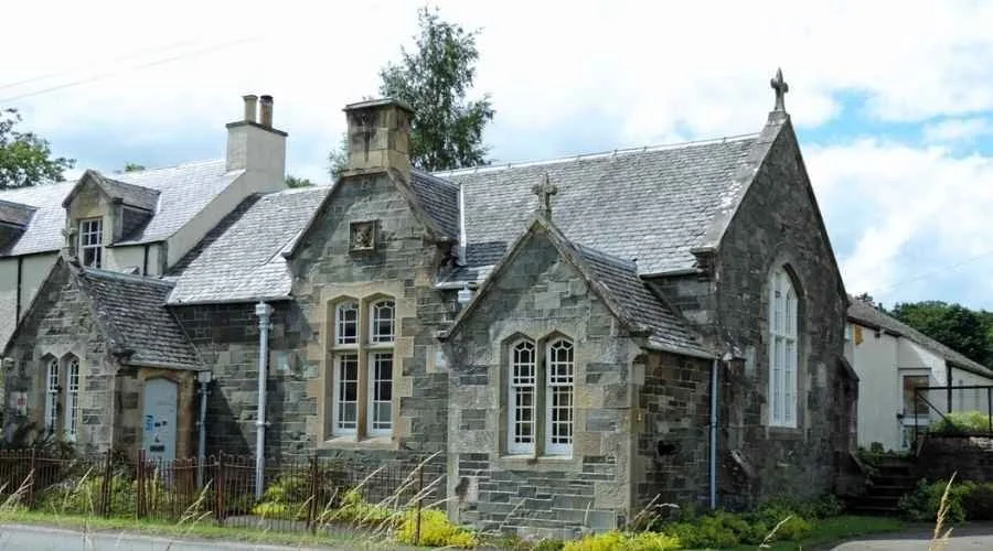 The 'Old School' building with a peaked roof and a cross on top, located in a rural area with a gravel path and grass in front.