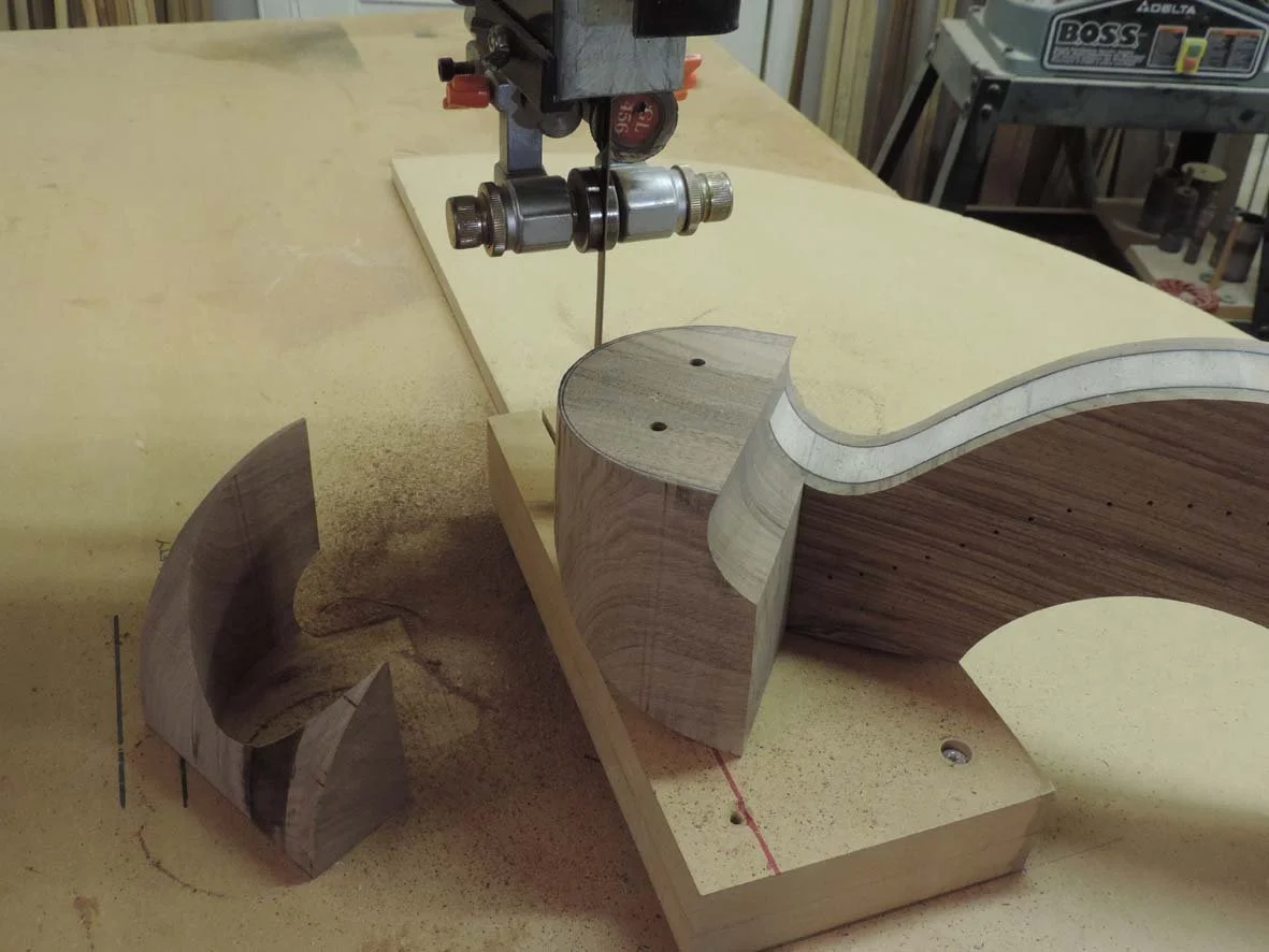 Woodworking workshop showing a partially assembled wooden harp on a workbench, with a clamp and a band saw in the background.