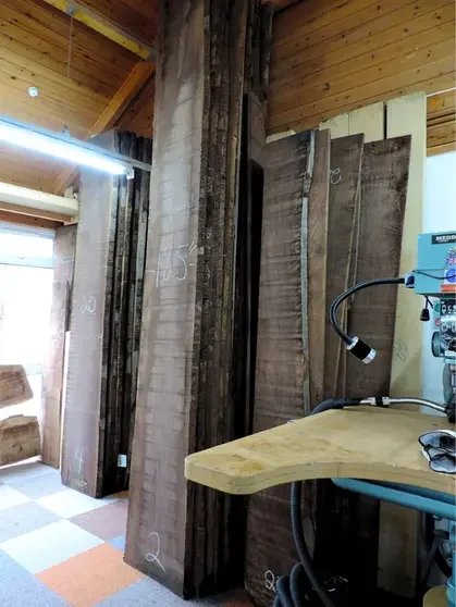 Stacked large wooden planks and beams in a workshop, with a wooden work table and various tools.