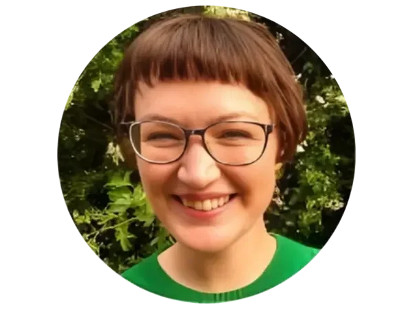 A woman with short brown hair, wearing glasses and a green shirt, smiling outdoors with green foliage in the background.