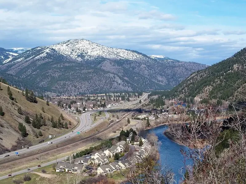 Mountain landscape with snow-capped peaks, a winding highway, a river, and a small town in a valley.