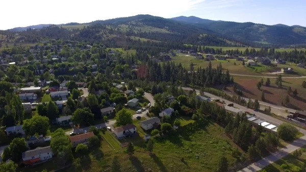 Aerial view of a rural town with houses, trees, and open fields surrounded by mountains in the distance.
